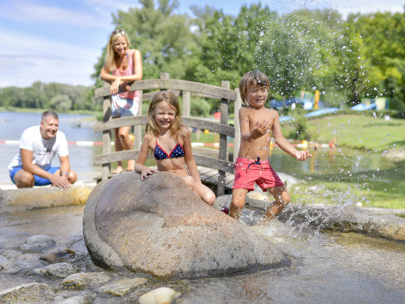 Kinder spielen bei einem großen Stein im Wasser Aubad Tulln, die Eltern schauen im Hintergrund zu, Tulln, Tullner Donauraum