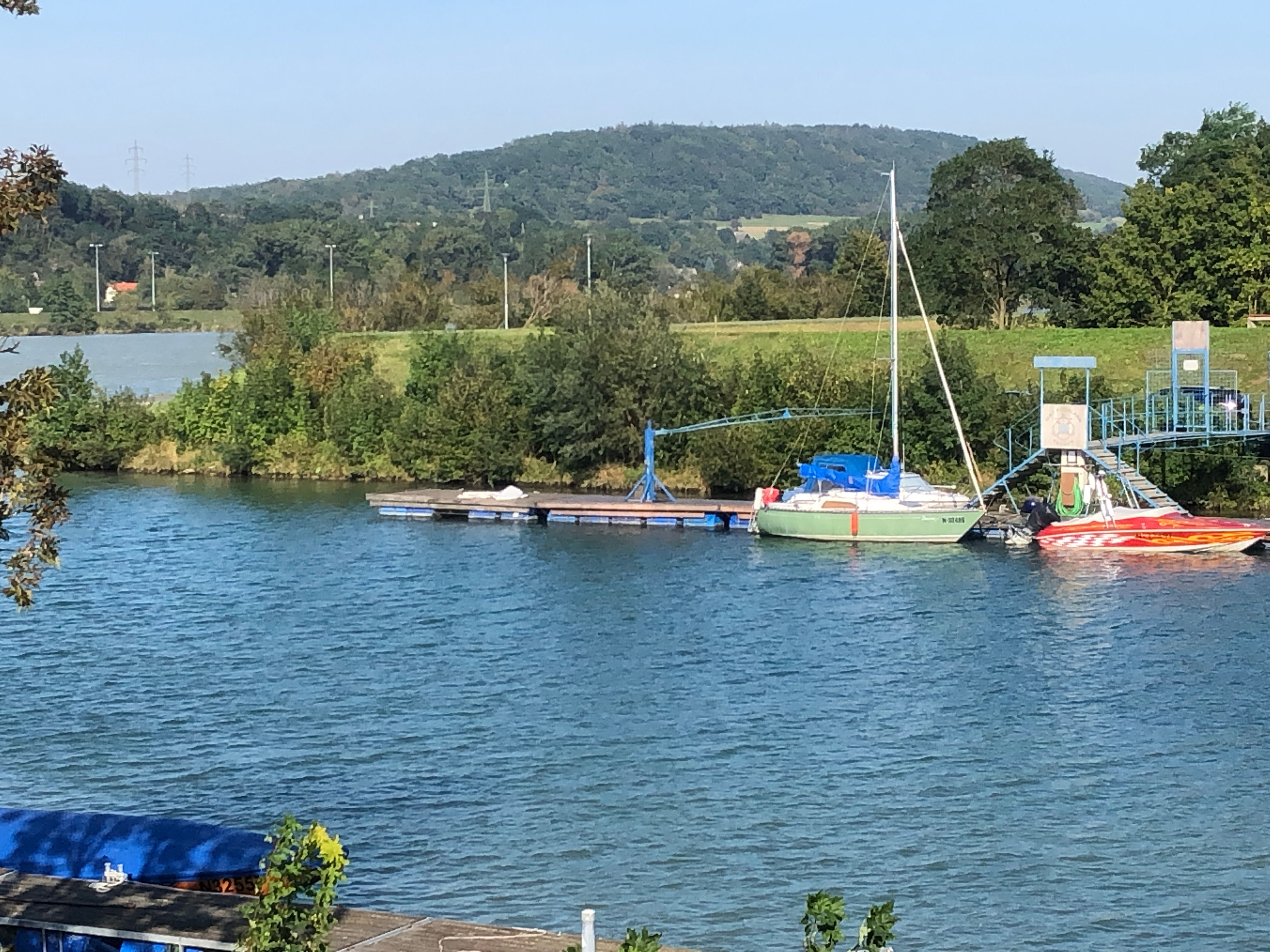 Boote an einem Steg auf einem Fluss mit bewaldeten Hügeln im Hintergrund.
