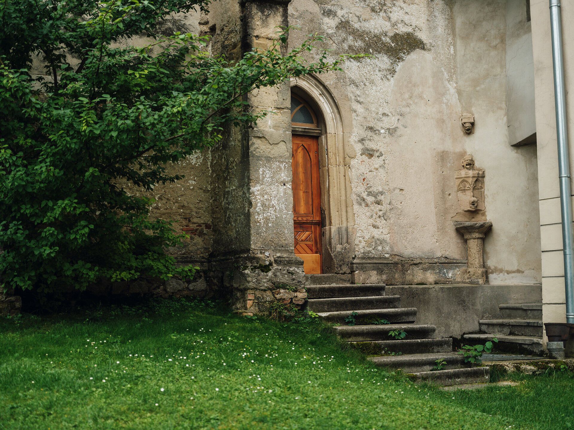 Seiteneingang einer Kirche mit Wiese und einem Baum davor, in der Ecke eine alte Skulptur, braune Türe mit Stiegen davor