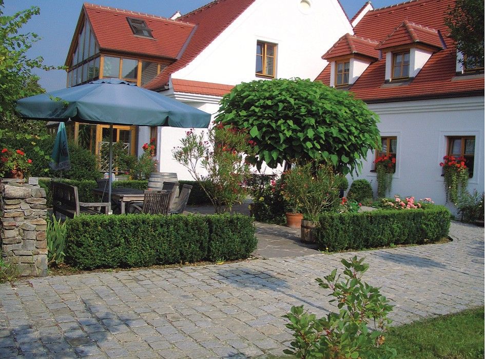 Ein gepflegter Garten mit Pflastersteinen, einem großen Sonnenschirm, Holzmöbeln und einem weißen Gebäude mit roten Dächern im Hintergrund.