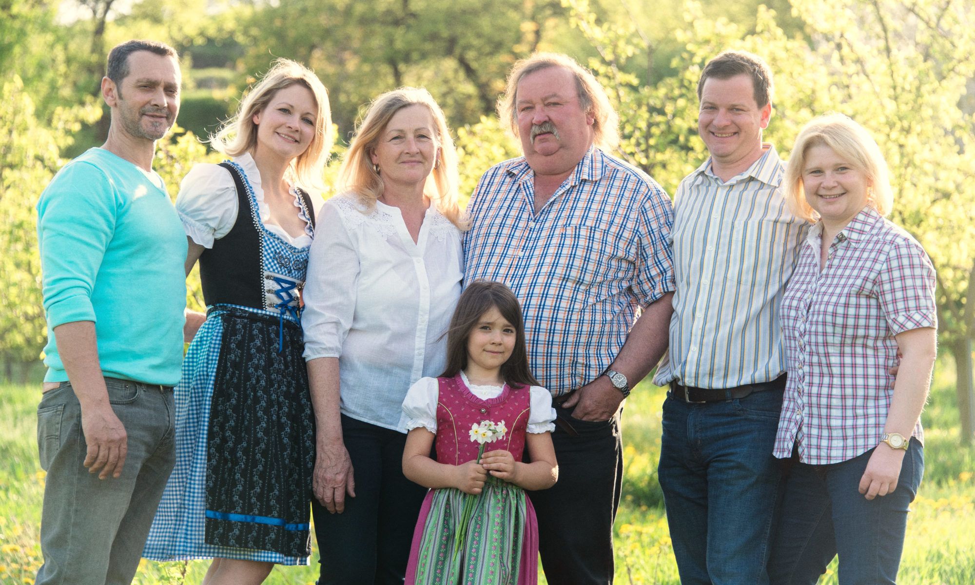 Eine Familie steht lächelnd in einem sonnigen Garten, einige tragen traditionelle Kleidung.