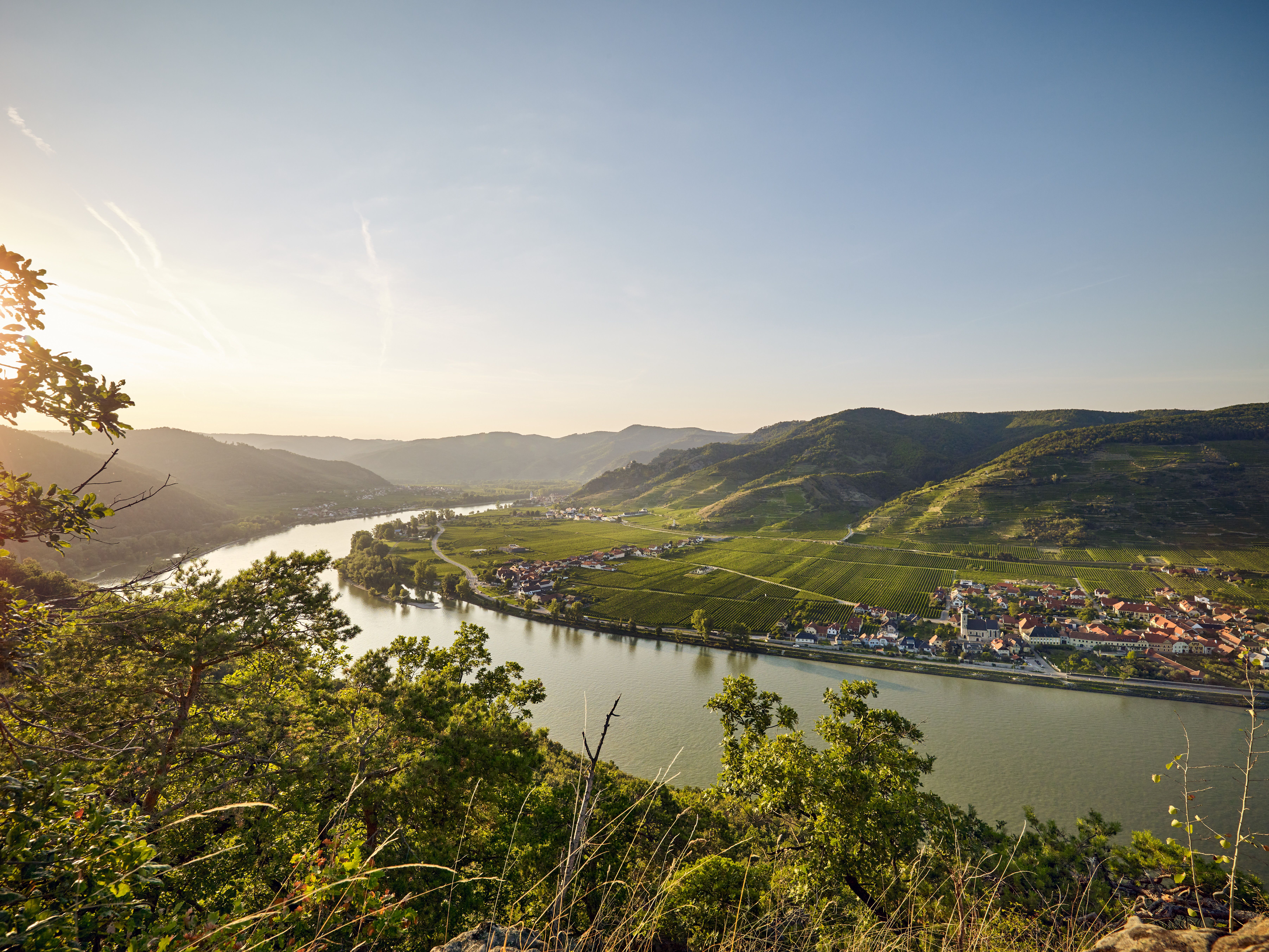 Panoramablick auf einen Fluss, umgeben von Hügeln und Dörfern, bei Sonnenuntergang.