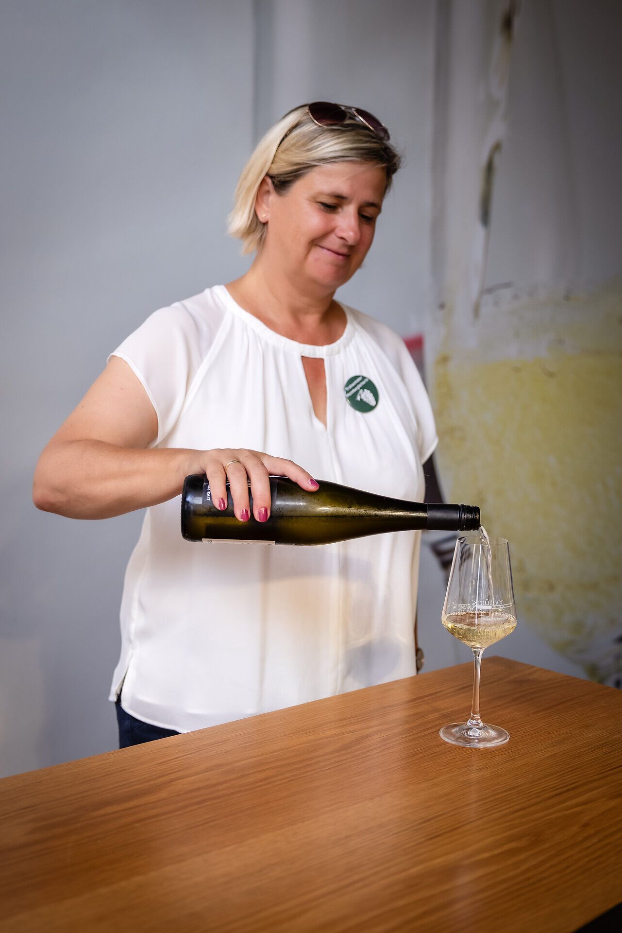 Blonde Frau schenkt Weißwein in ein Glas, welches auf einem Holztisch steht