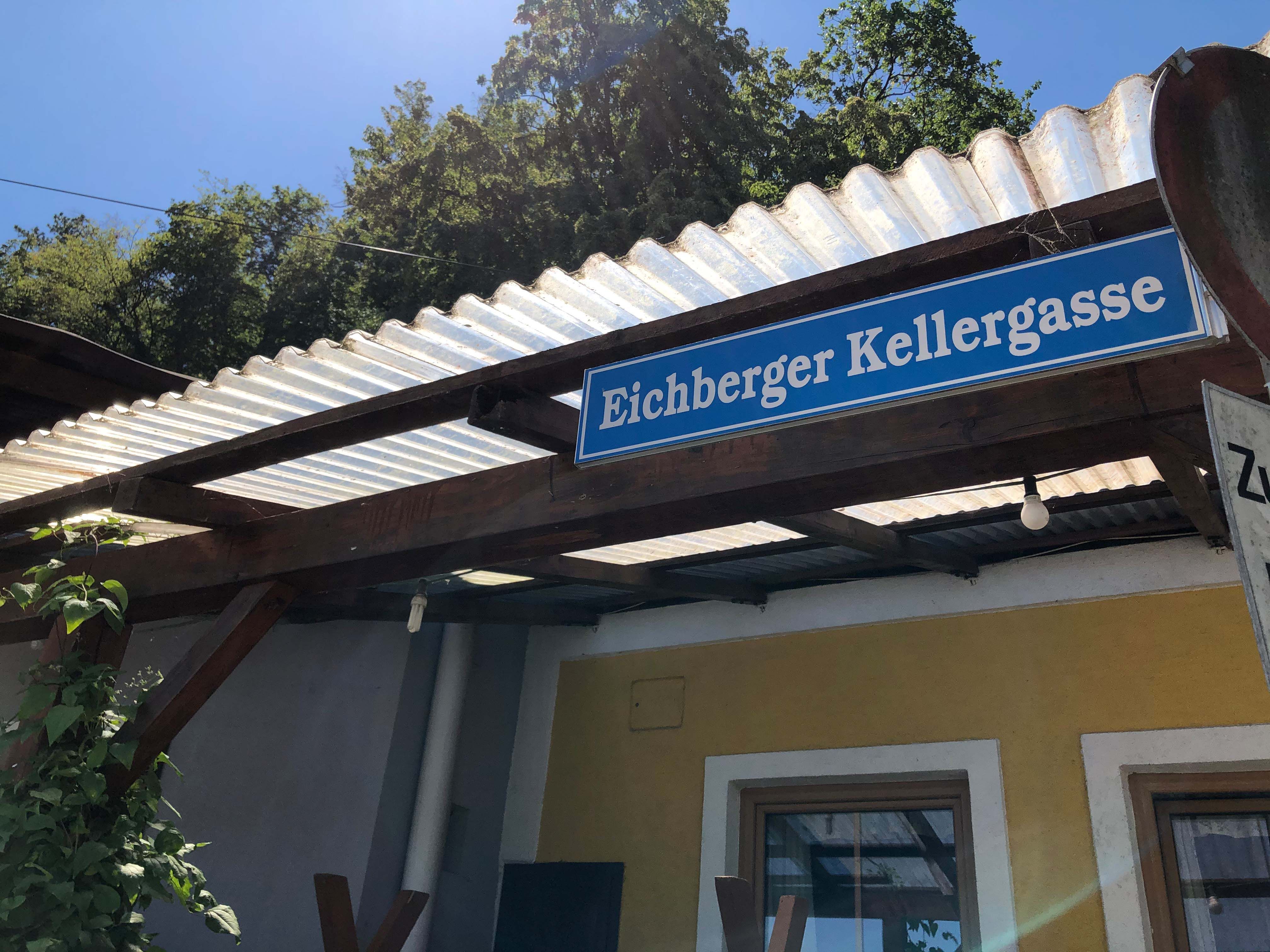 Schild mit der Aufschrift 'Eichberger Kellergasse' an einem Gebäude mit Wellblechdach.
