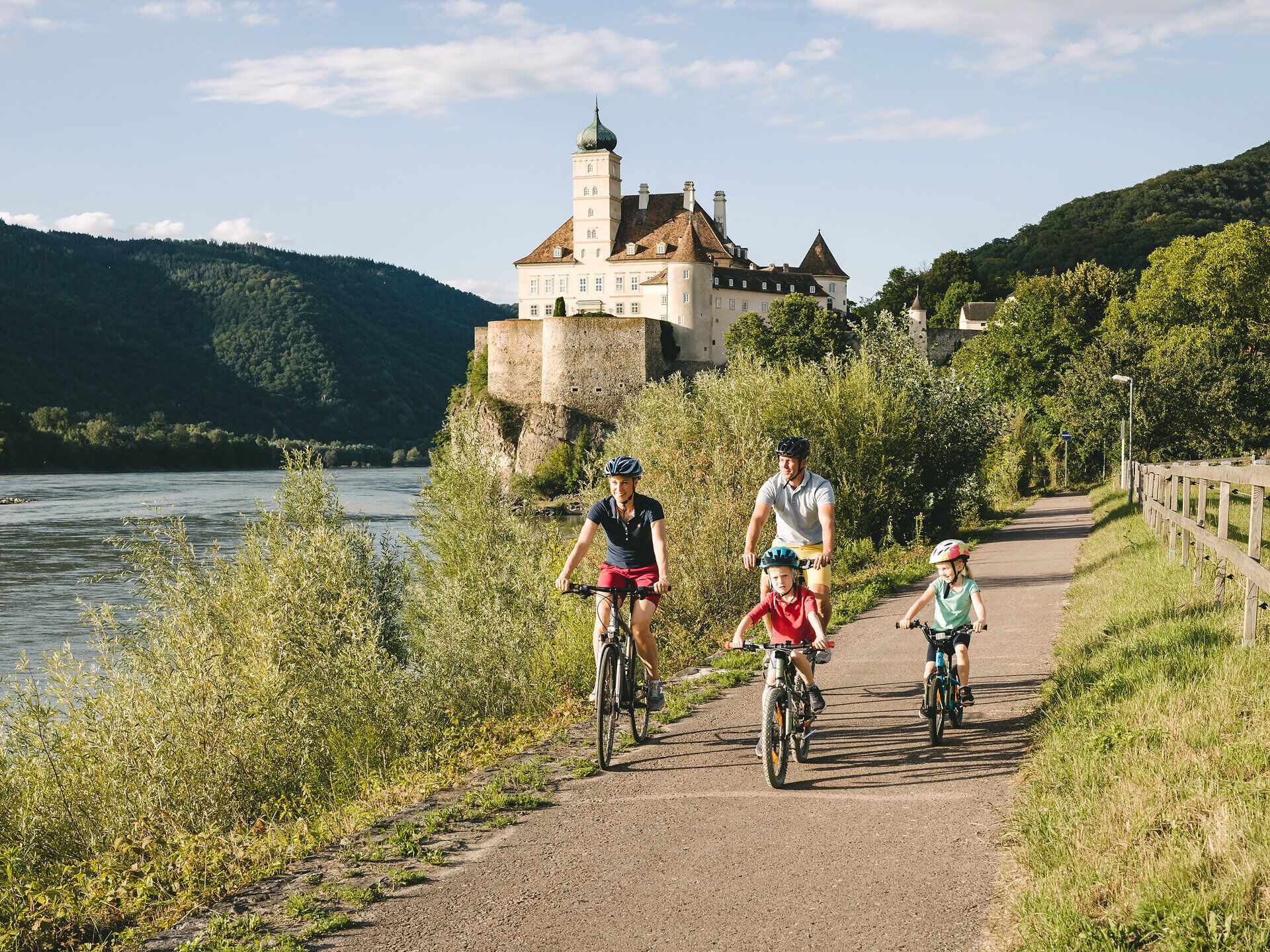 Eine Familie genießt einen sonnigen Tag am Radweg, umgeben von üppigem Grün und der malerischen Kulisse eines majestätischen Schlosses. Die Kinder lachen und strampeln fröhlich auf ihren Fahrrädern, während der Fluss sanft plätschert und die Berge im Hintergrund eine friedliche Atmosphäre schaffen.