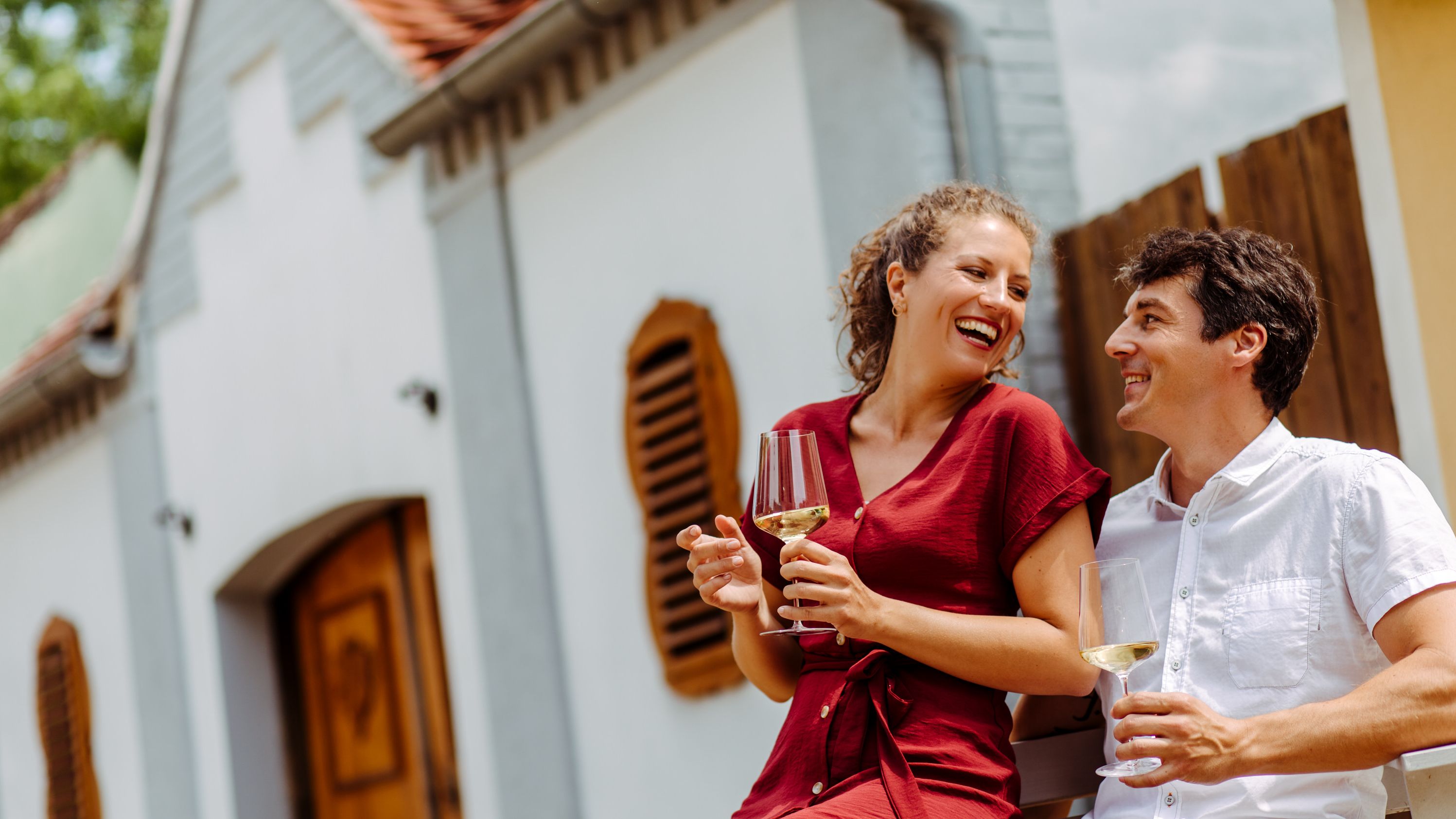 Zwei Personen sitzen vor einem Weinkeller und halten Gläser mit Weißwein.