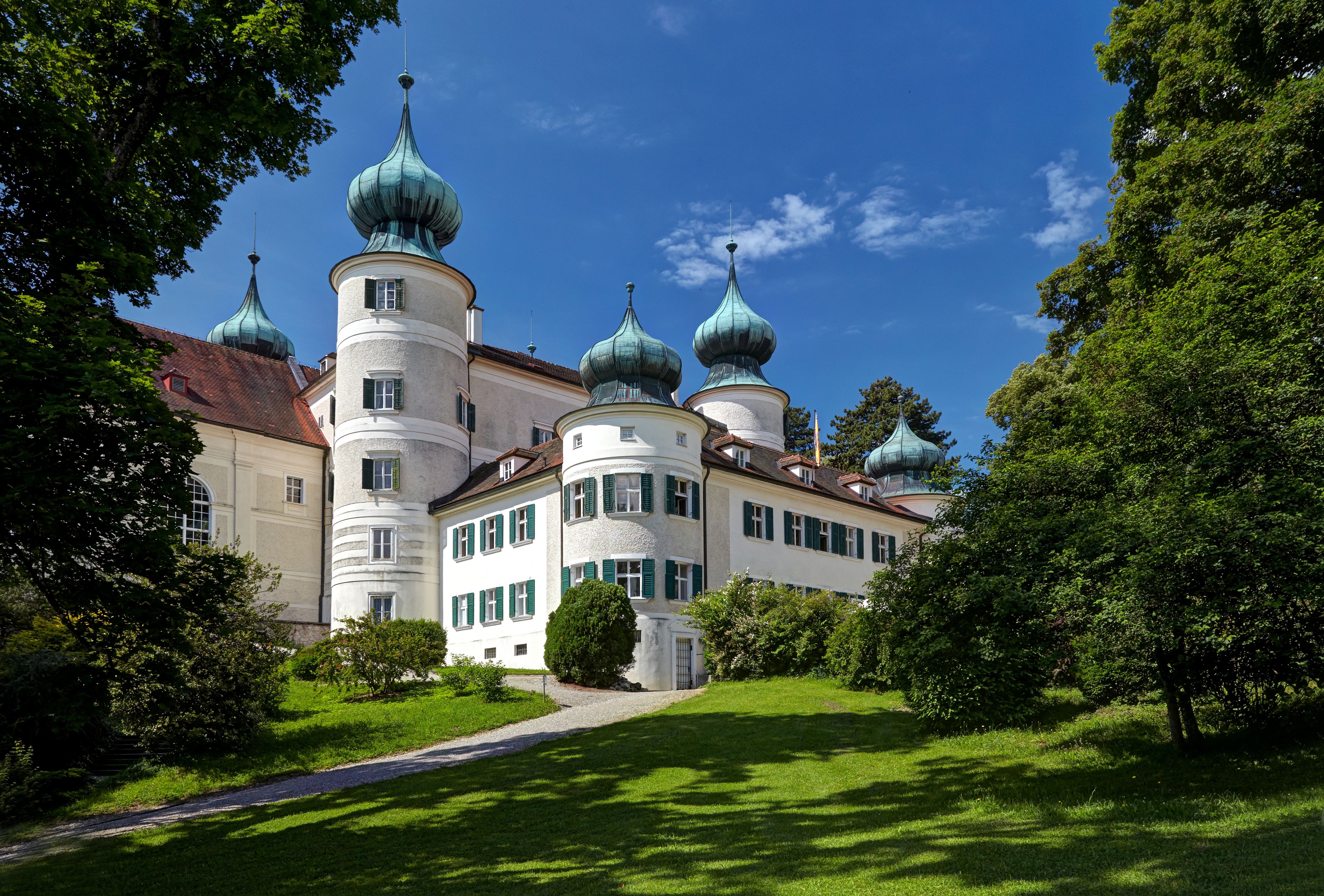 A white castle with several onion domes, surrounded by green countryside and trees, under a blue sky.