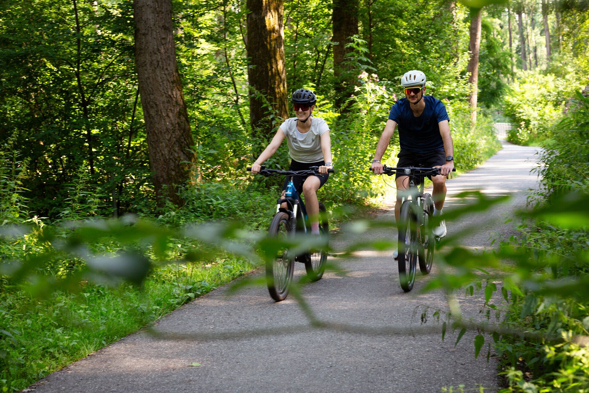 Zwei Radfahrer fahren auf einem Weg umgeben von Bäumen und grünen Sträuchern.