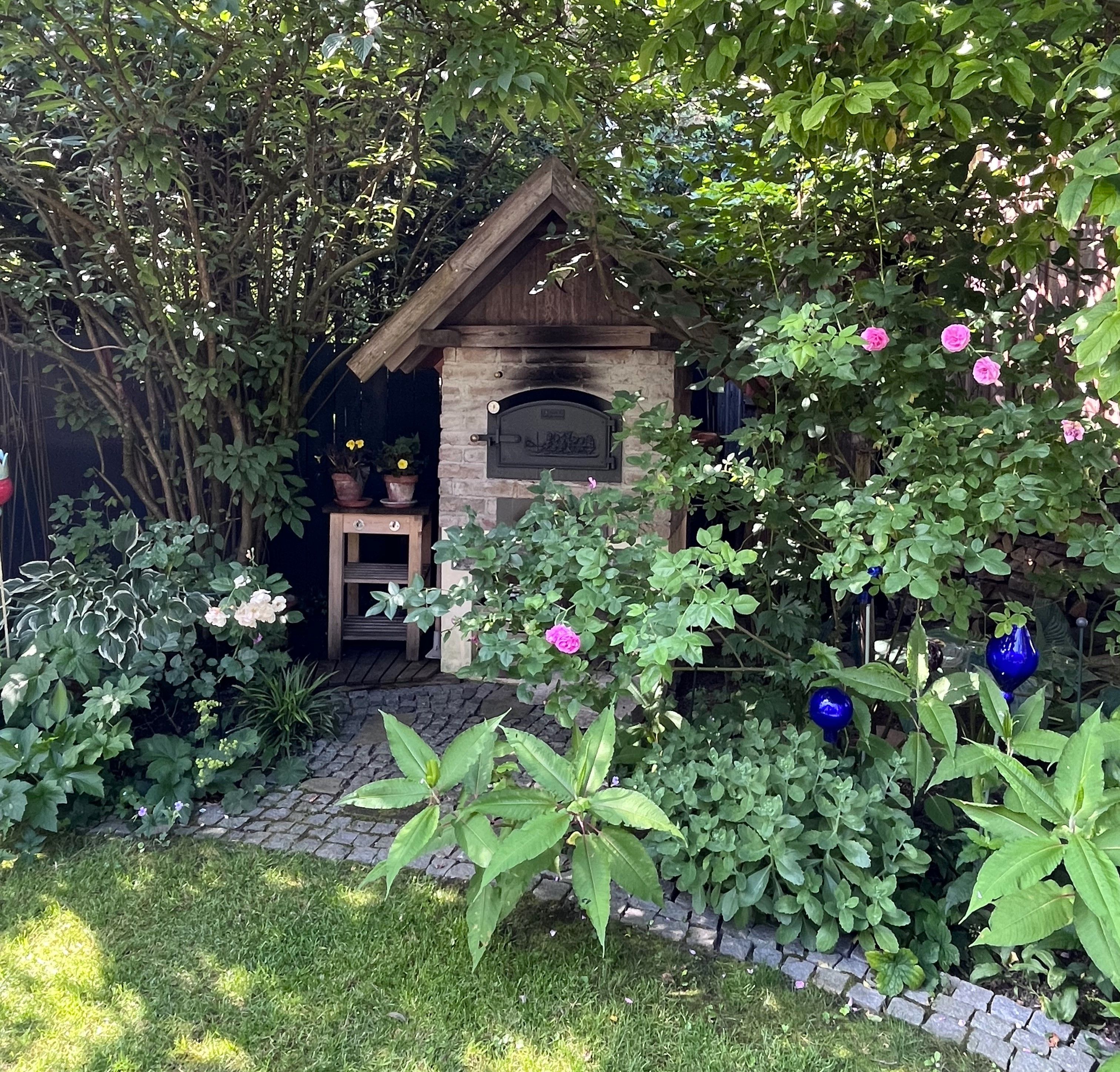 Ein gemauerter Ofen in einem üppigen Garten mit blühenden Rosen und grünen Pflanzen.