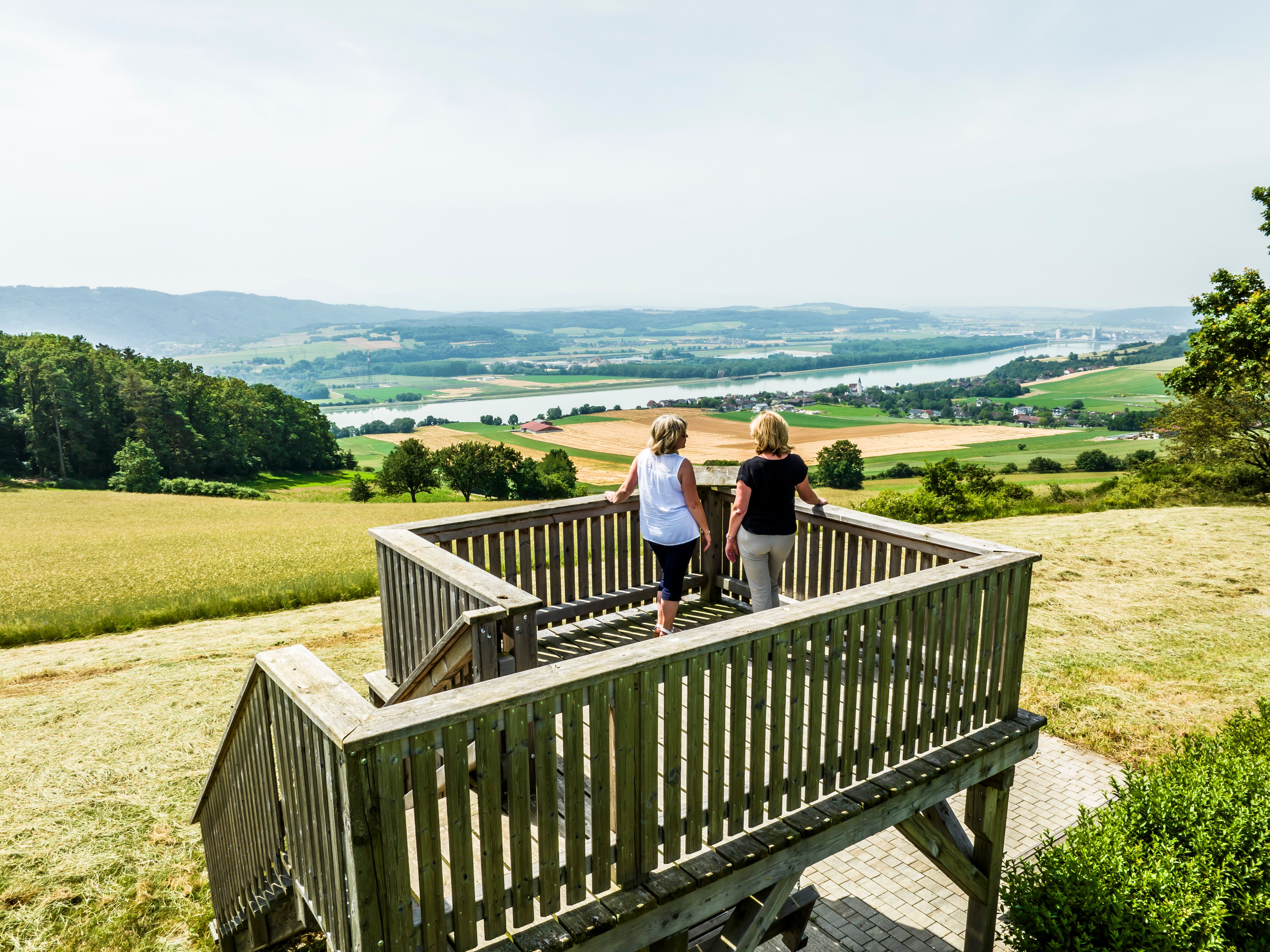 Two people stand on a wooden viewing platform with a view of a wide landscape and a river.