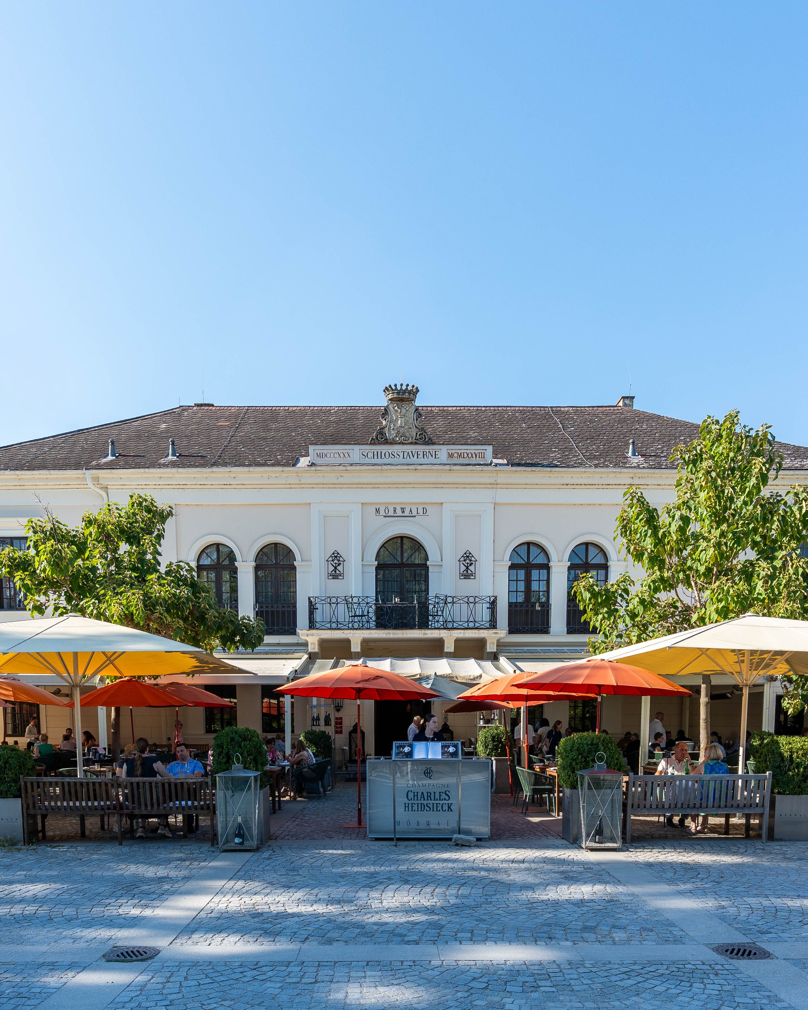 Pohled zepředu na Grafenegg Hotel & Restaurant s terasou, slunečníky a hosty před budovou Mörwald v Grafeneggu.