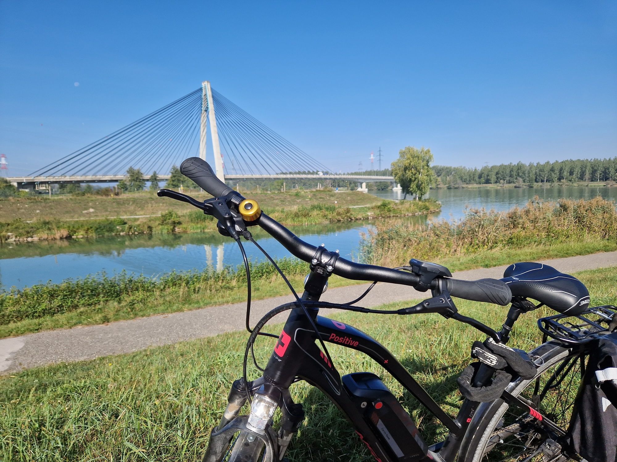 Ein E-Bike steht vor einer modernen Brücke über einen Fluss, umgeben von grüner Landschaft und blauem Himmel.