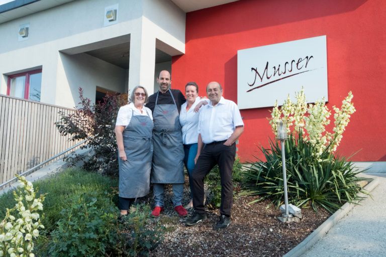 Vier Personen stehen vor einem Gebäude mit einem Schild 'Musser'.