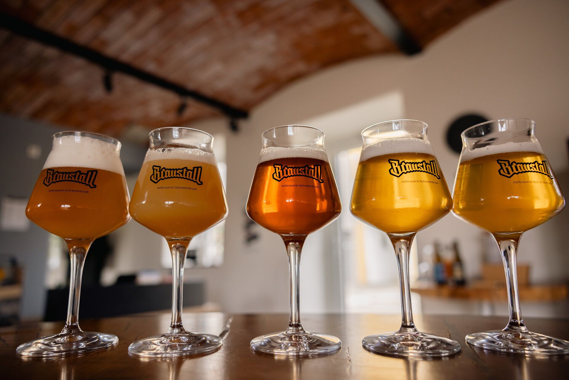 Fünf gefüllte Biergläser mit Braustall-Logo in unterschiedlichen Bierfarben auf einer Holztheke.