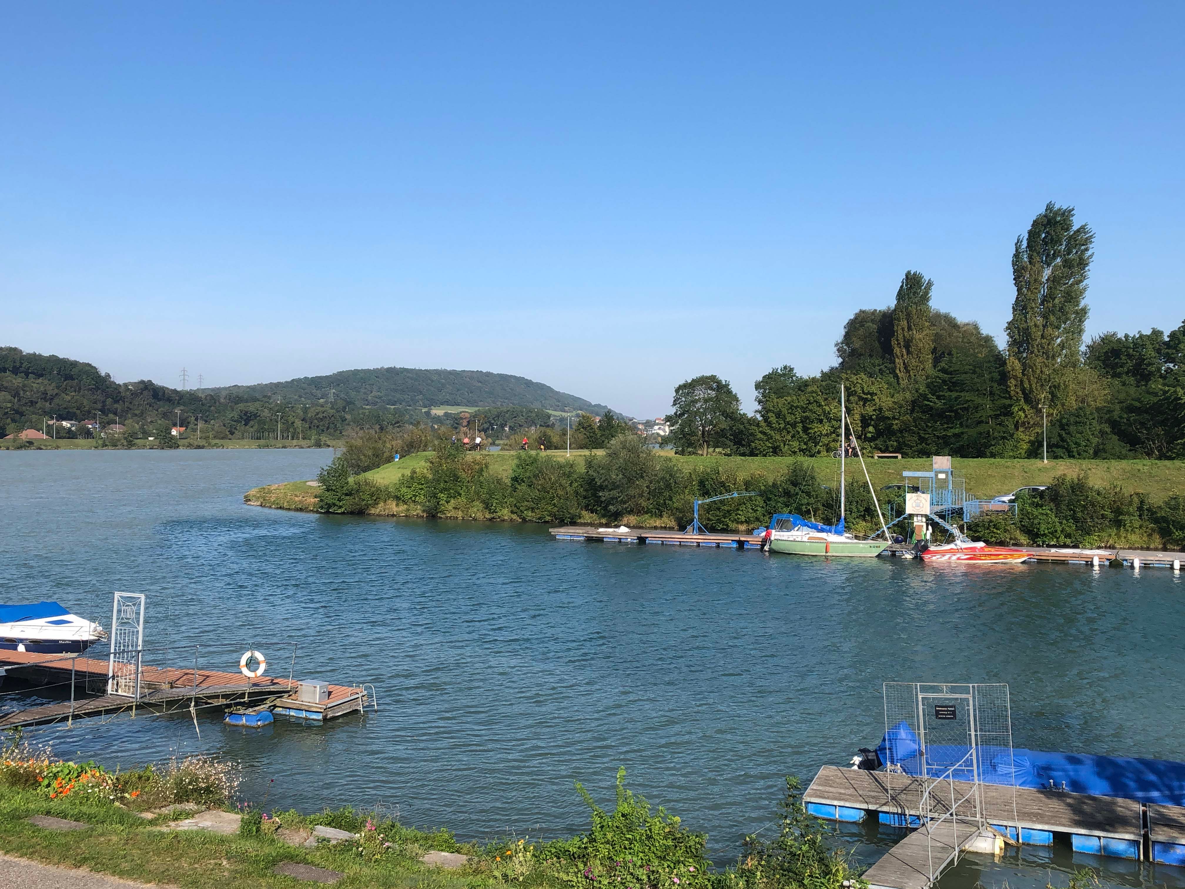 Anlegestelle mit Booten am Flussufer, umgeben von grüner Landschaft und blauem Himmel.