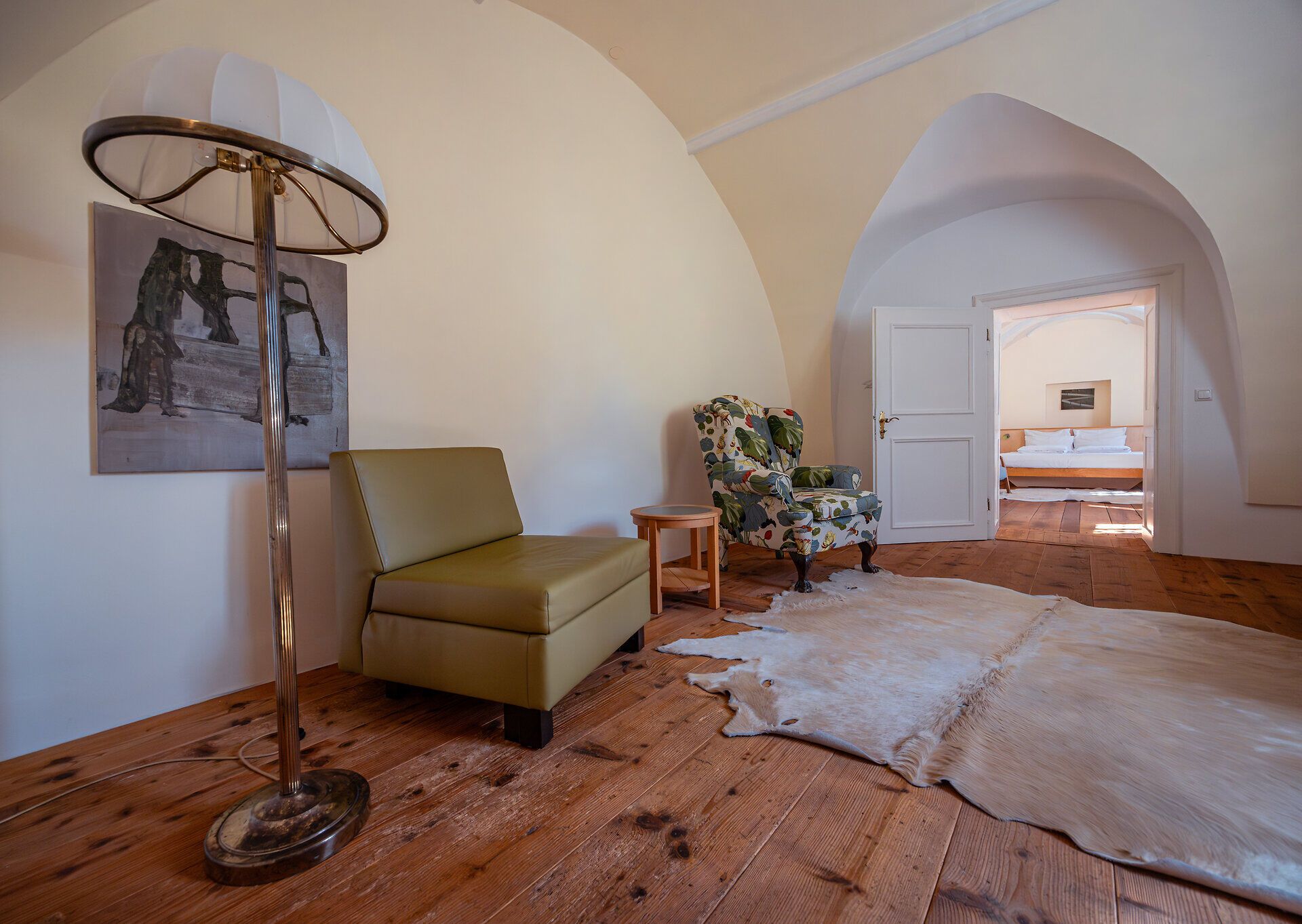 Ein einladender Raum mit warmen Holzböden und sanften Farben schafft eine entspannte Atmosphäre. Die stilvolle Einrichtung, bestehend aus einem bequemen Sessel und einer modernen Stehlampe, lädt zum Verweilen ein. Ein weicher Teppich aus Tierfell ergänzt das gemütliche Ambiente und verspricht einen angenehmen Aufenthalt.