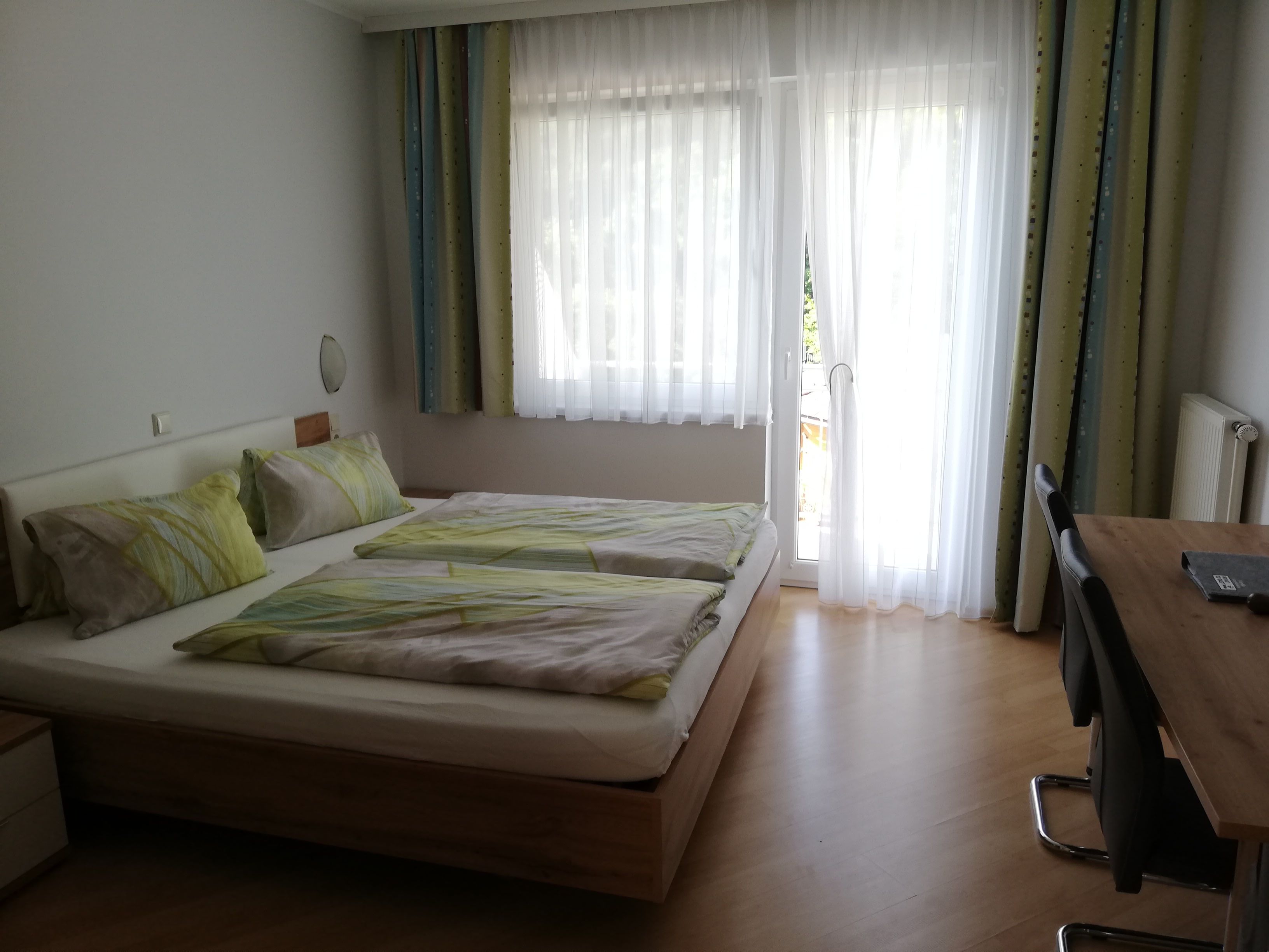 Ein Doppelzimmer mit einem Bett, einem Tisch und einem Balkon mit Vorhängen.
