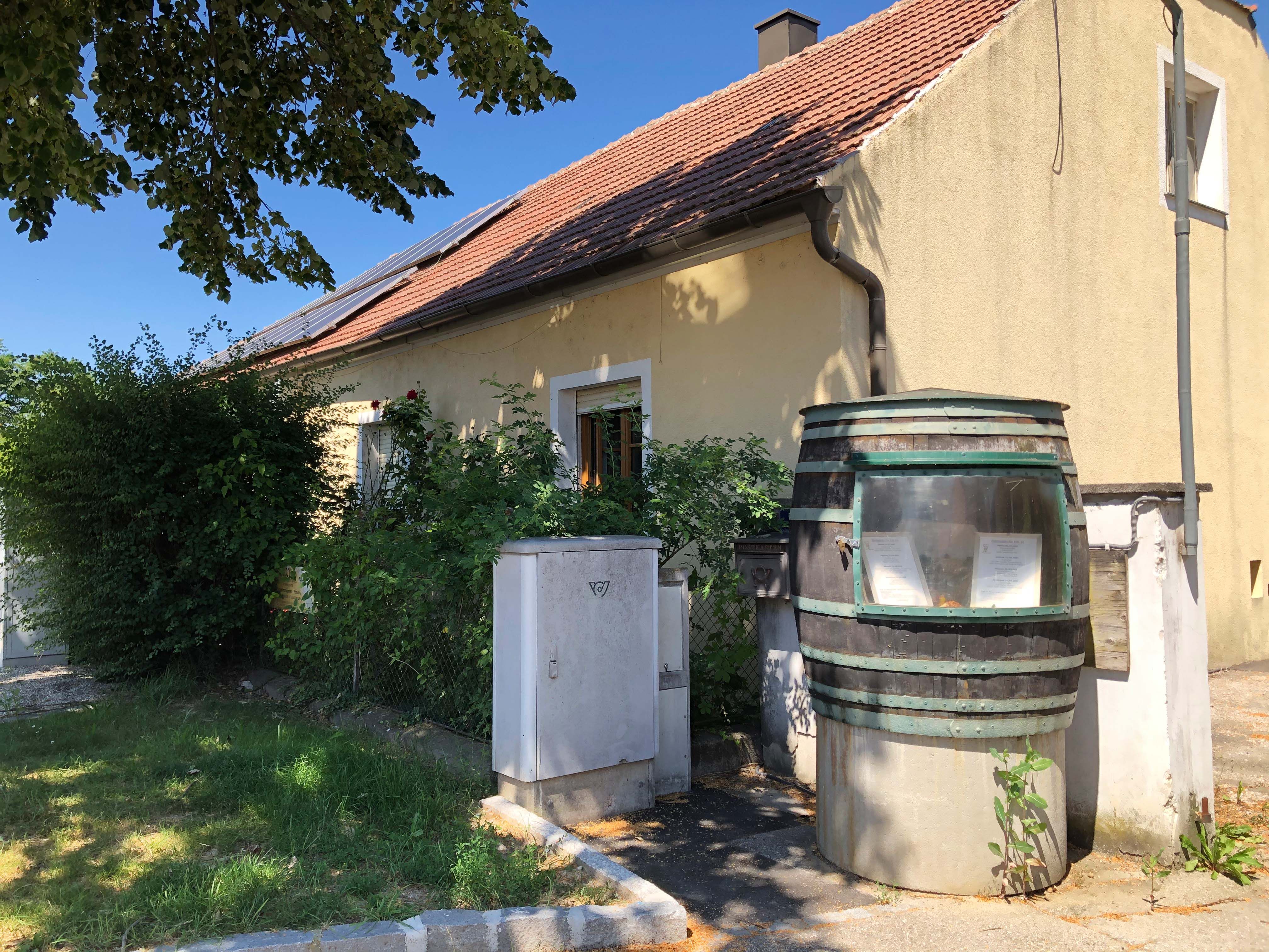 Ein gelbes Haus mit rotem Ziegeldach und Solarpanelen. Im Vordergrund ein großes Fass mit Schauglas und Pflanzen.