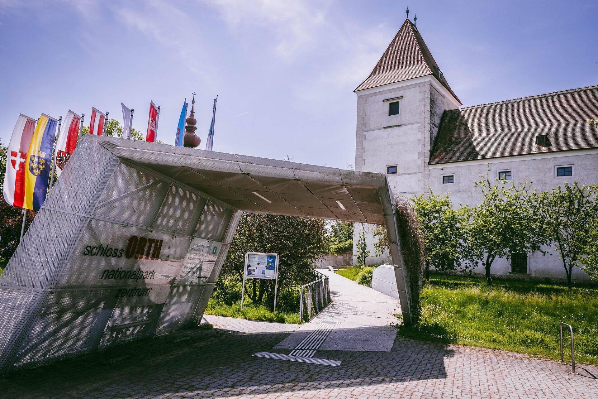 Das beeindruckende Schloss Orth thront majestätisch über der Donau und lädt Besucher ein, die Schönheit der umliegenden Natur zu entdecken. Die bunten Fahnen, die im sanften Wind wehen, verleihen dem Ort eine festliche Atmosphäre und versprechen unvergessliche Erlebnisse im Nationalpark Donau-Auen.