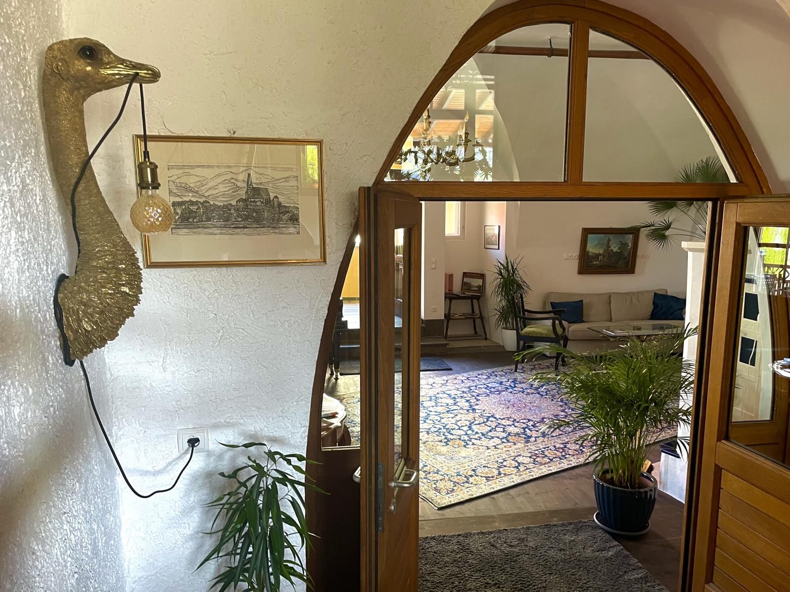 Innenansicht eines Wohnzimmers mit Pflanzen, einem Sofa und einem dekorativen Wandobjekt in Form eines Vogels mit einer Lampe.