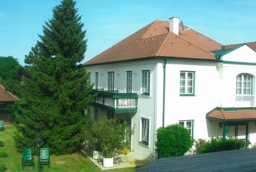 Ein zweistöckiges weißes Gästehaus mit rotem Dach und grünen Fensterläden, umgeben von einem Garten mit Bäumen und Gartenmöbeln.
