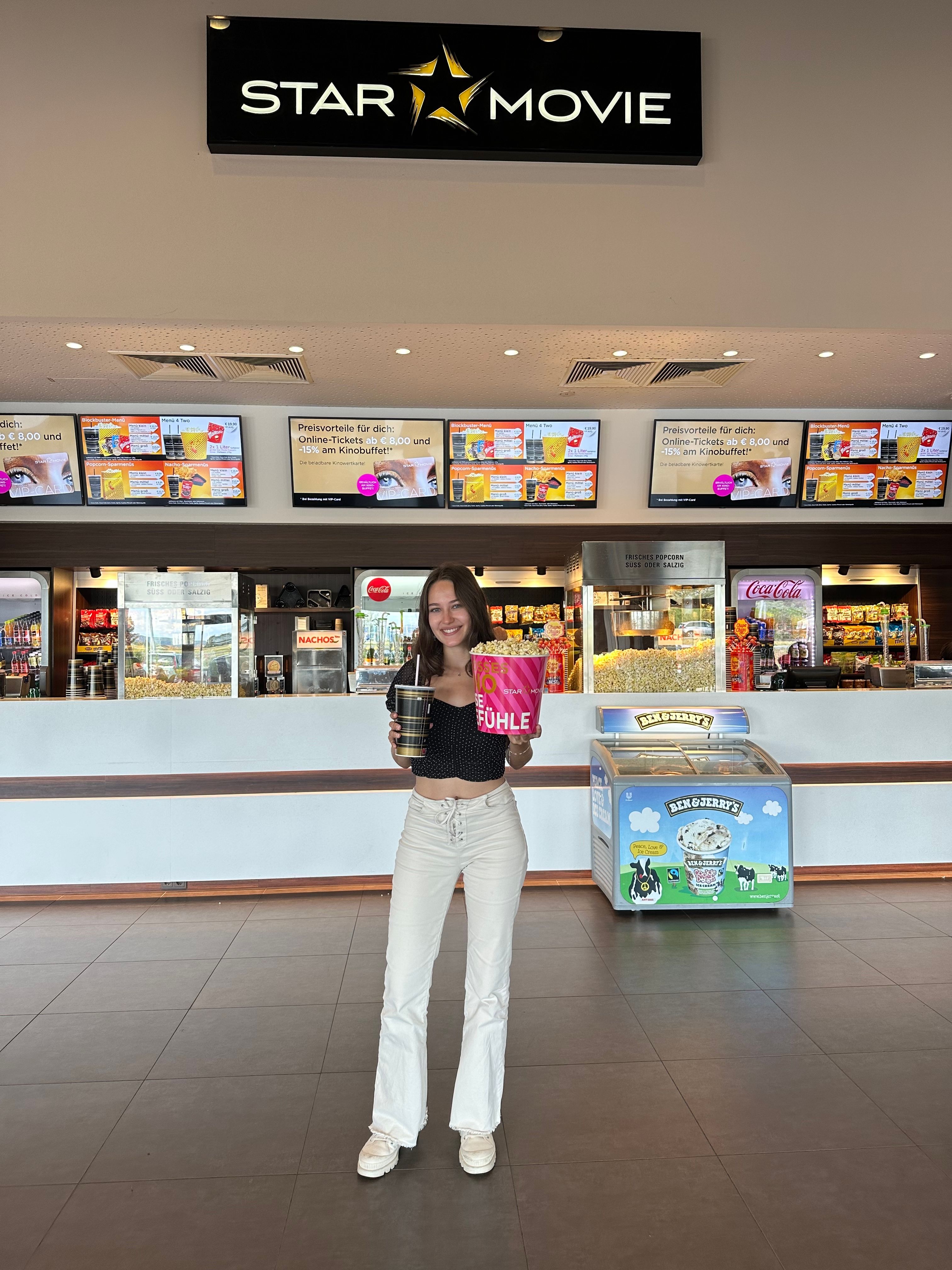 Eine Frau steht in einem Kinofoyer vor einem Star Movie Schild mit Popcorn und Getränk in den Händen.