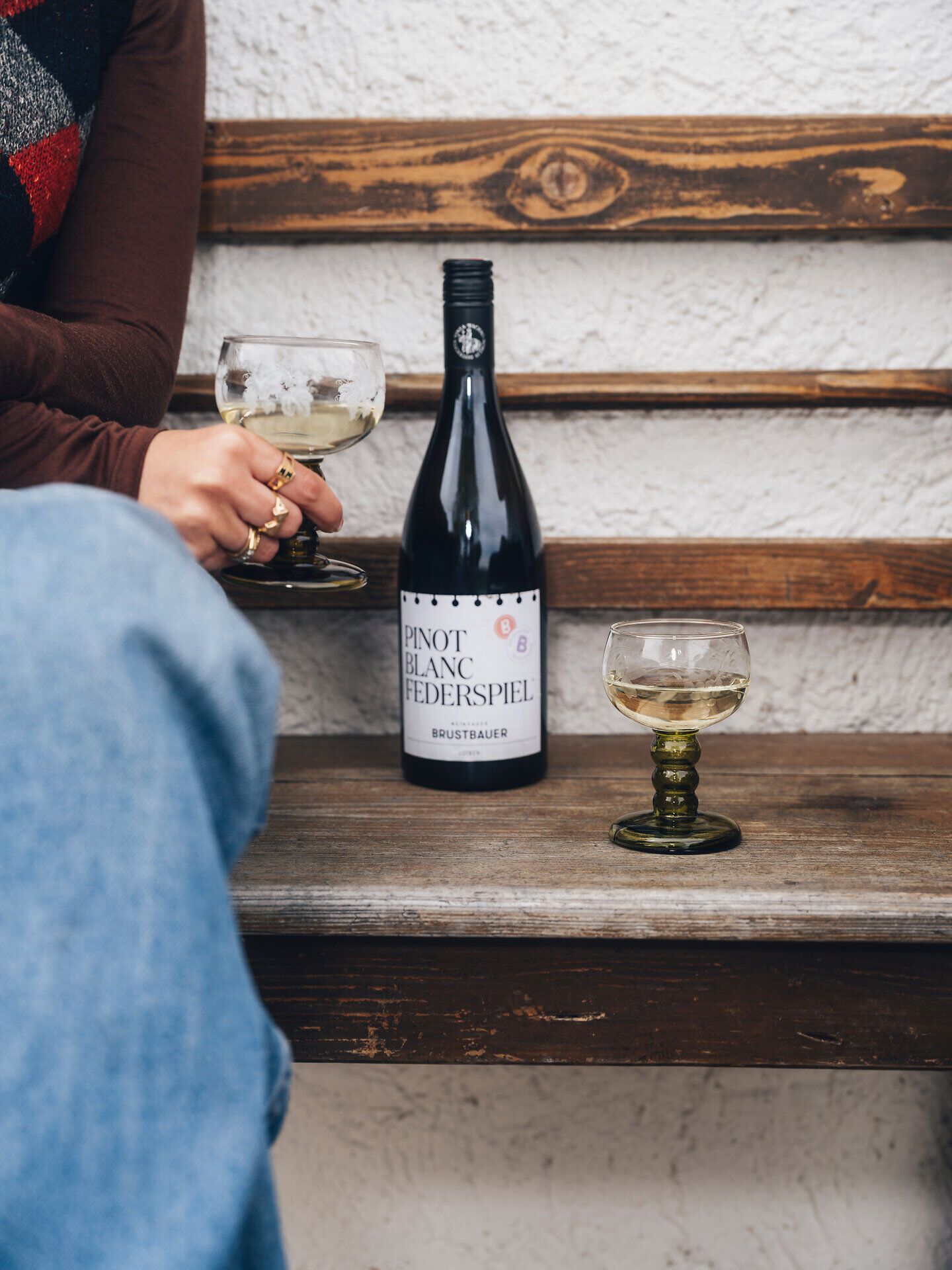 Weinflasche steht auf einer Holzbank neben zwei Gläsern mit Weißwein; eine Person sitzt daneben, sichtbar sind Jeans und Hand mit Glas.