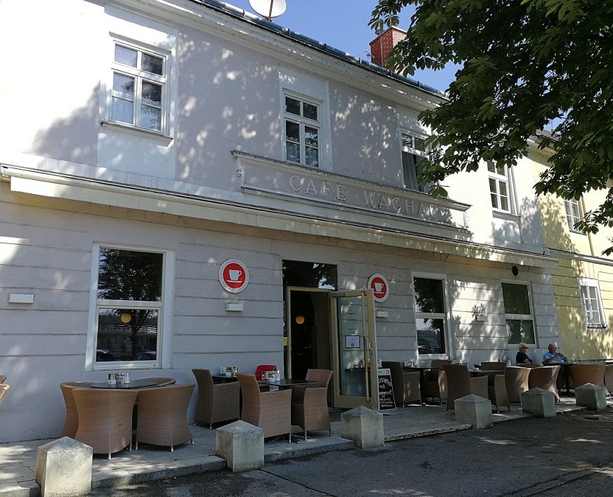 Außenansicht eines Cafés mit Terrasse und Sitzgelegenheiten unter einem Baum.
