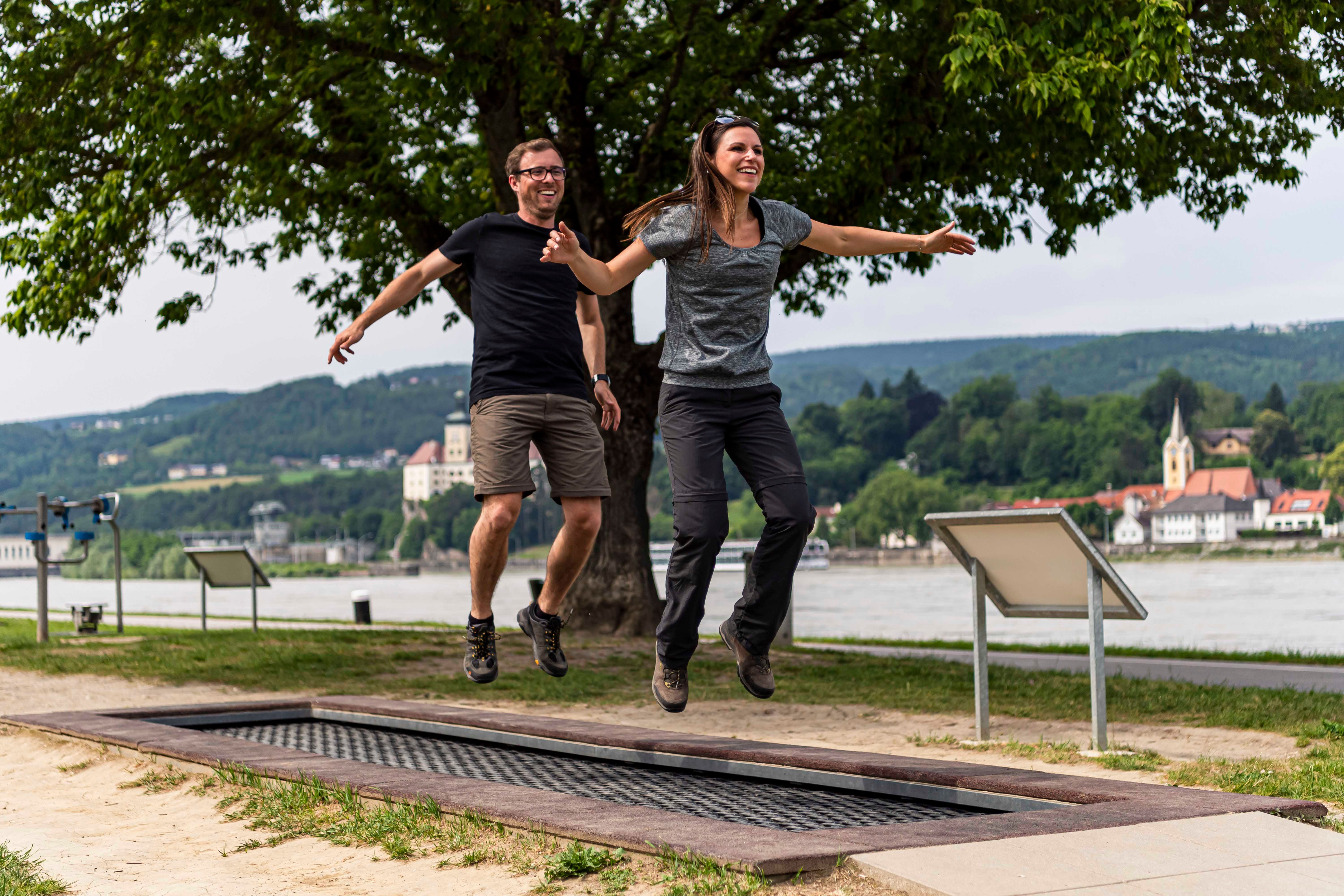 Two people jumping on a trampoline outdoors, with a river and buildings in the background.