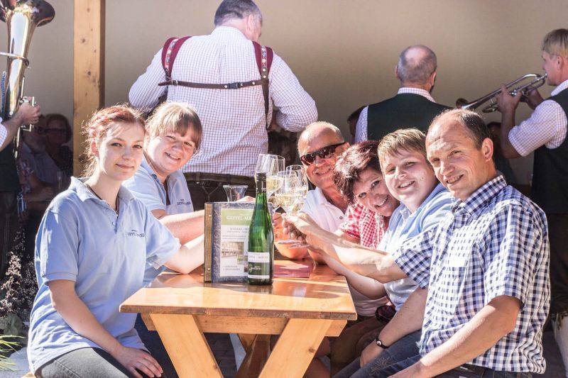Gruppe von Menschen sitzt an einem Tisch im Freien und stößt mit Getränken an.