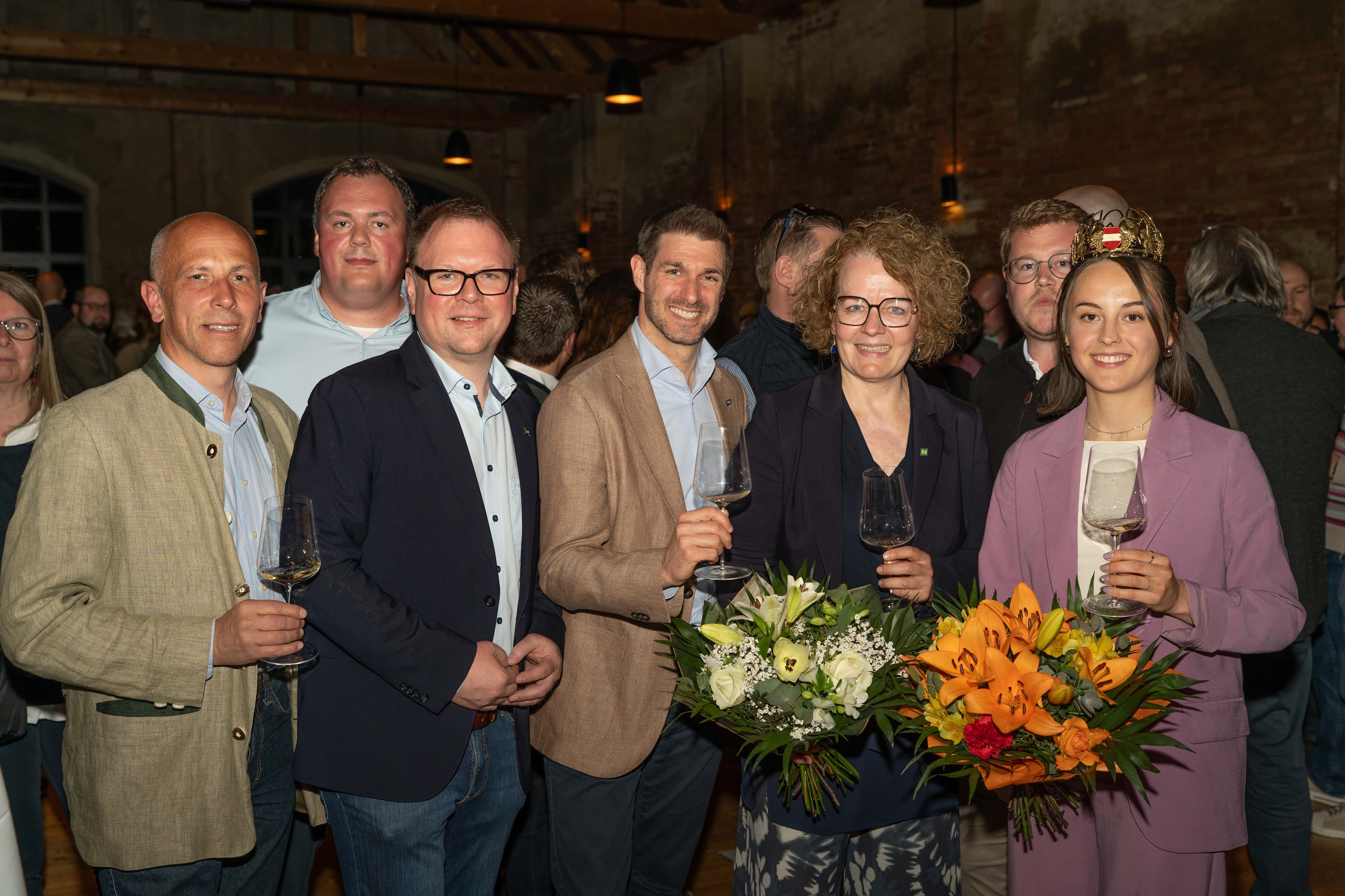 Mehrere Gäste stehen zusammen bei einer Weinveranstaltung in einer rustikalen Eventlocation mit Backsteinwänden, halten Weingläser und Blumensträuße und posieren für ein gemeinsames Foto während der Feier.
