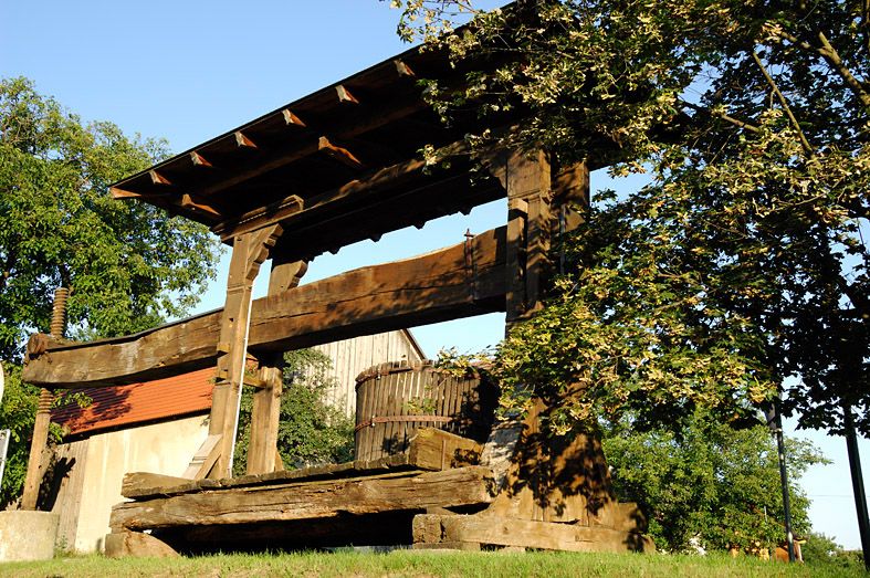 Alte hölzerne Weinpresse im Freien, umgeben von Bäumen und Gebäuden.