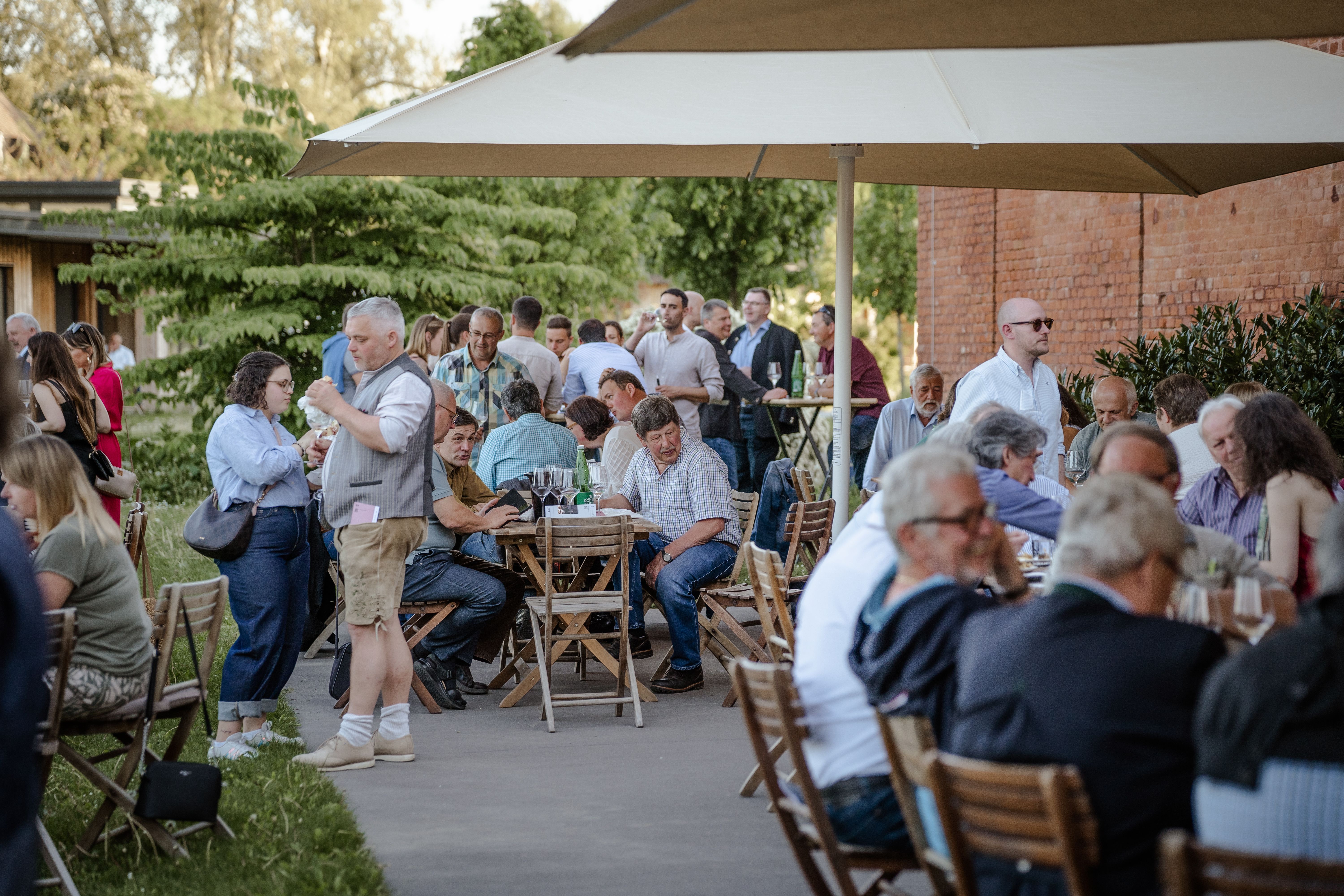 Viele Menschen sitzen dicht beieinander an Tischen im Freien vor der alten Scheune -  eine gesellige Stimmung unter Bäumen bei Tageslicht.