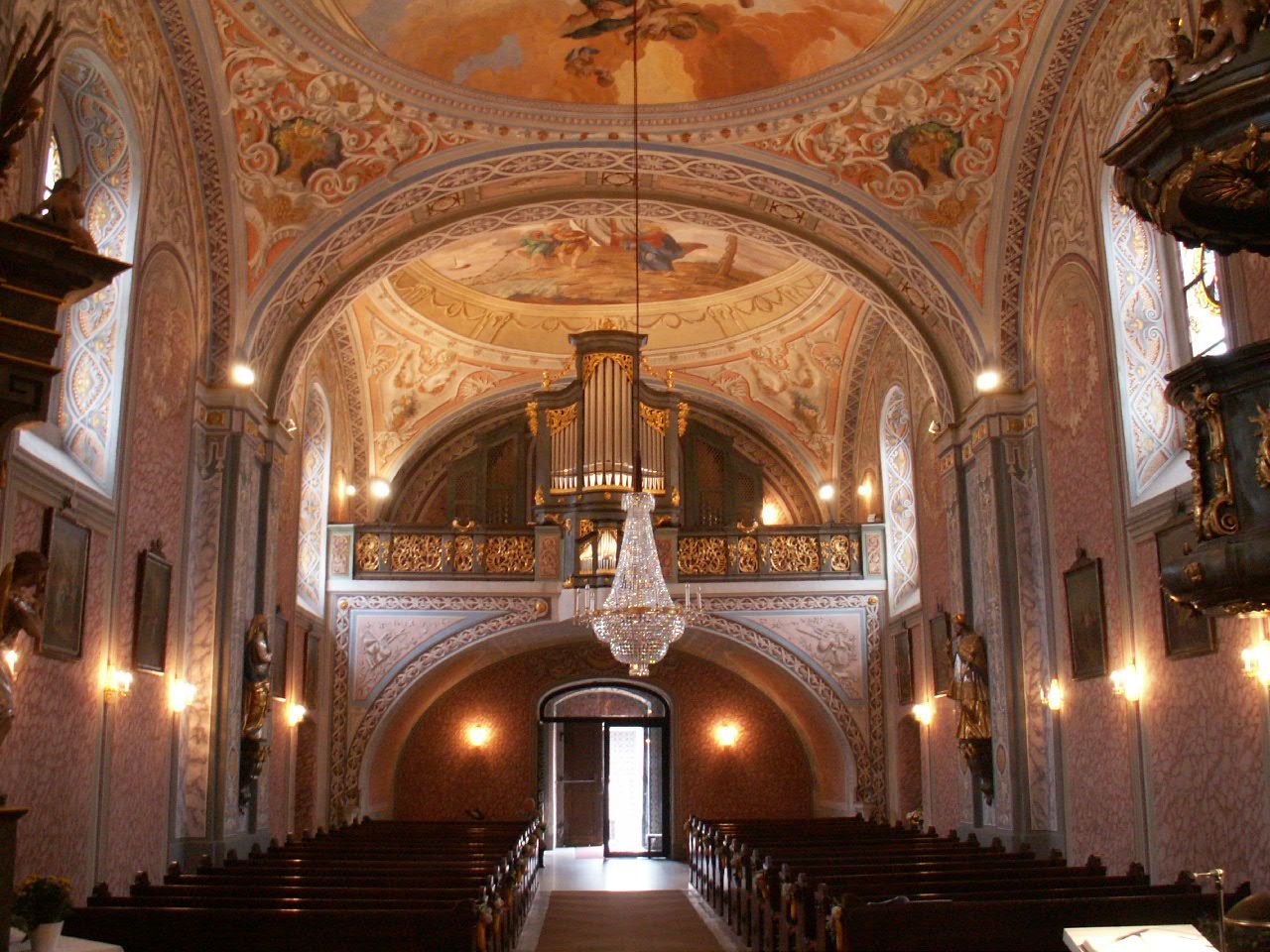 Blick vom Altarraum durch das Kirchenschiff einer barocken Kirche mit Deckenfresken, Orgelempore, Kronleuchter und hölzernen Kirchenbänken.