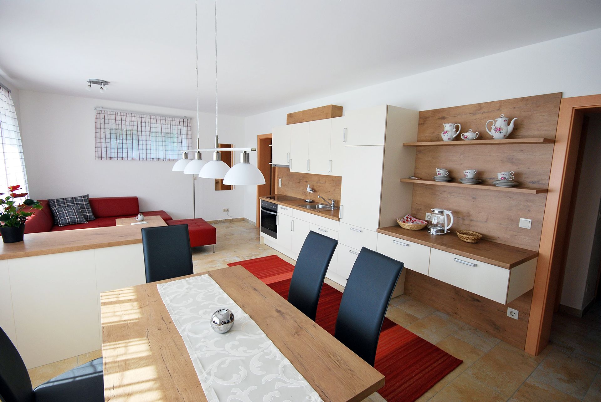 Moderner Wohn- und Essbereich mit offener Küche, rotem Sofa, Holztisch und weißen Hängelampen.