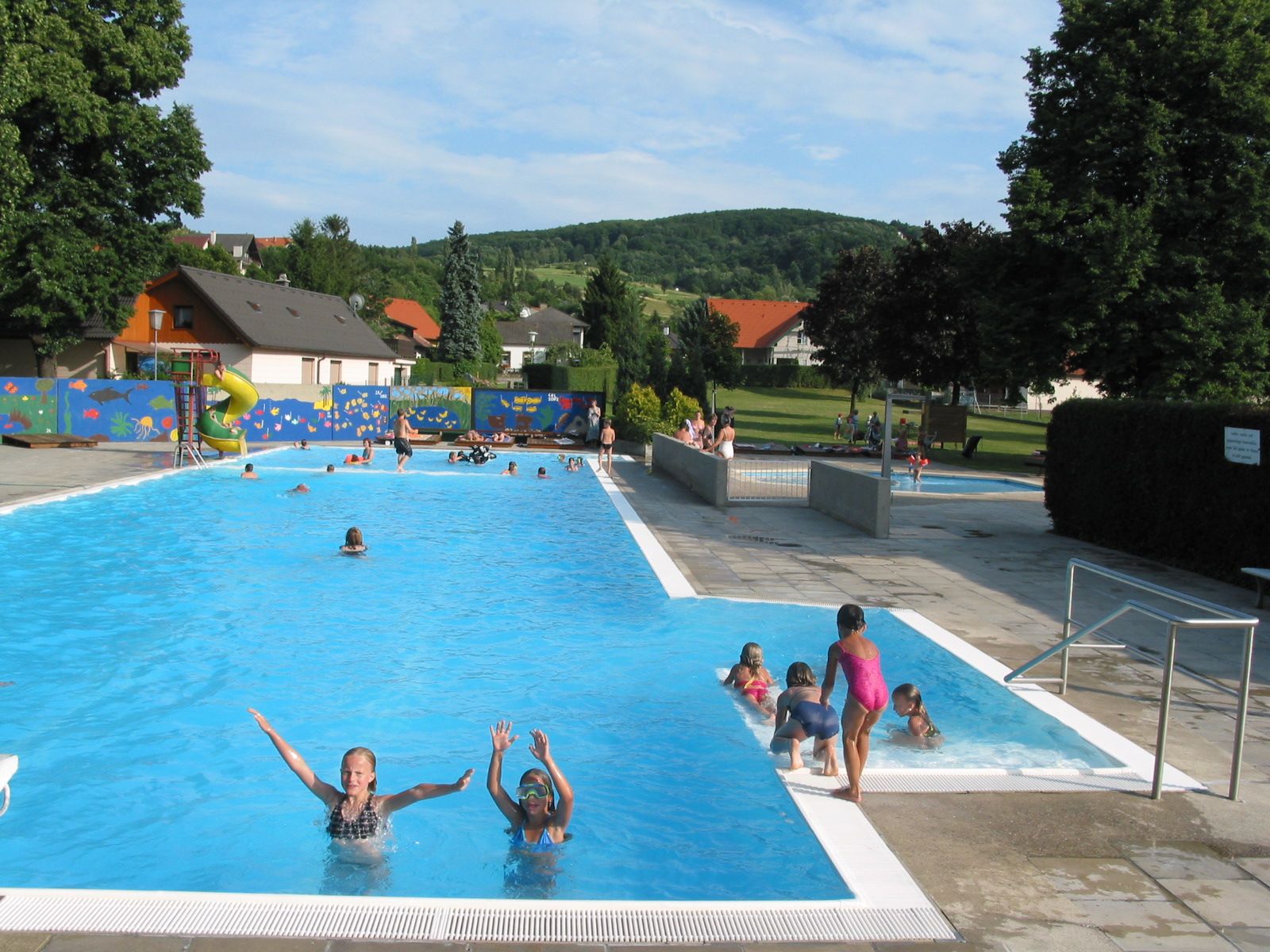 Freibad mit Schwimmern und Rutsche, umgeben von Bäumen und Wiese.