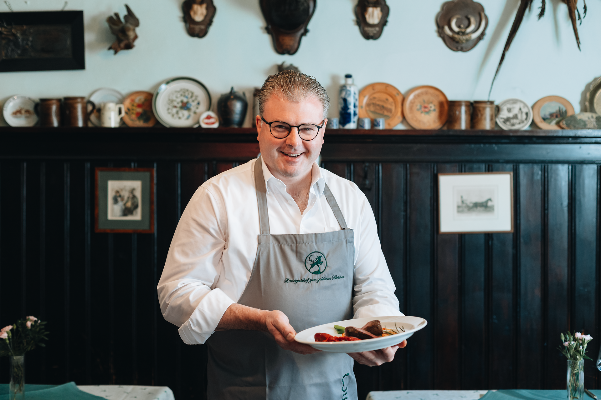 In einem einladenden Wirtshaus, umgeben von traditioneller Wirtshauskultur, präsentiert der Wirt stolz ein kunstvoll angerichtetes Gericht. Die warmen Farben und die rustikale Atmosphäre laden dazu ein, die köstlichen Speisen und die herzliche Gastfreundschaft zu genießen.