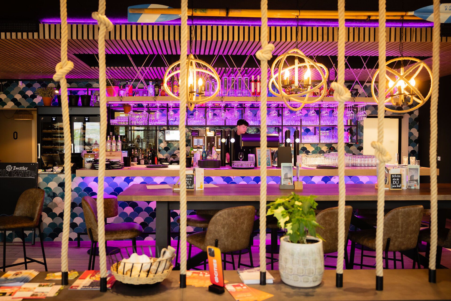 Blick durch Seile auf eine beleuchtete Bar mit einem Barkeeper