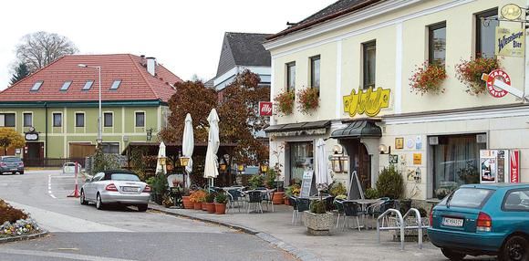 Exterior view of an inn on a lively square with trees, parasols, armchairs and much more.