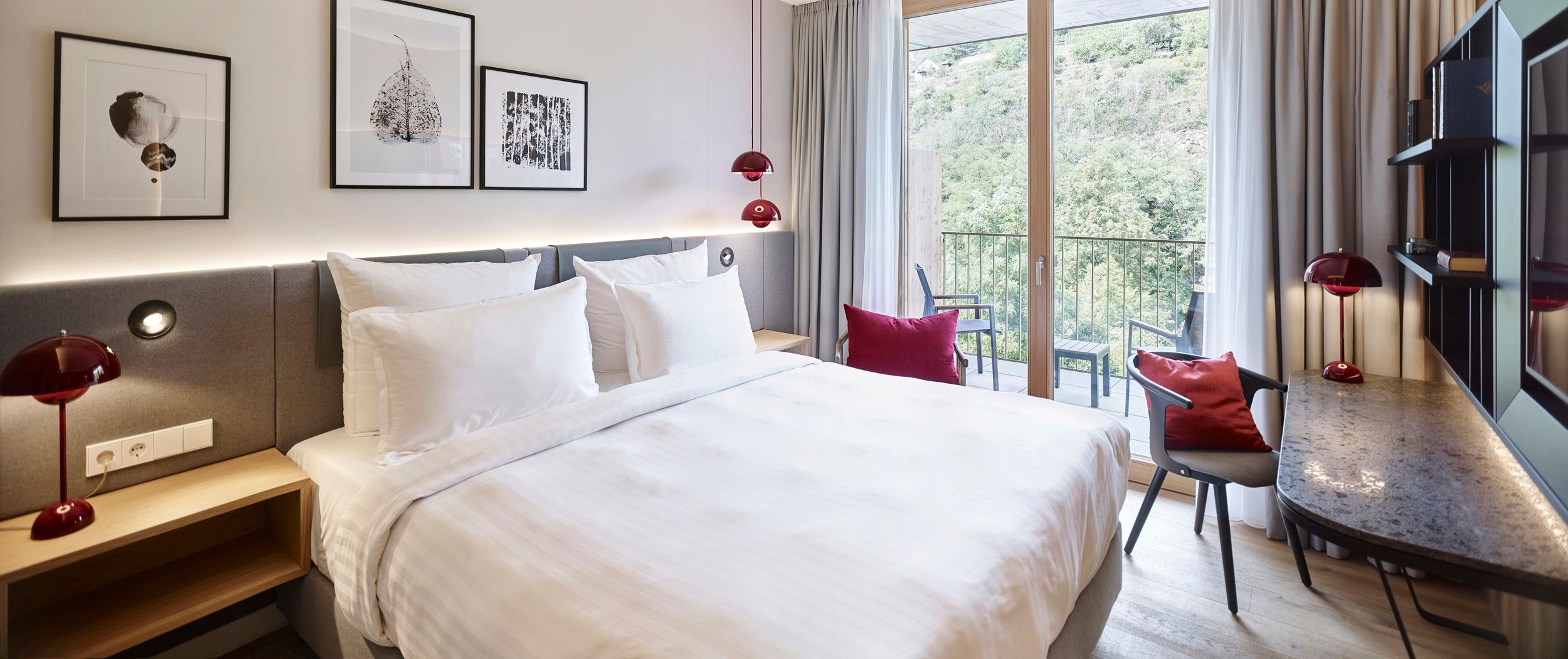 Modernes Hotelzimmer mit großem Bett, Schreibtisch und Balkon mit Aussicht auf grüne Landschaft.