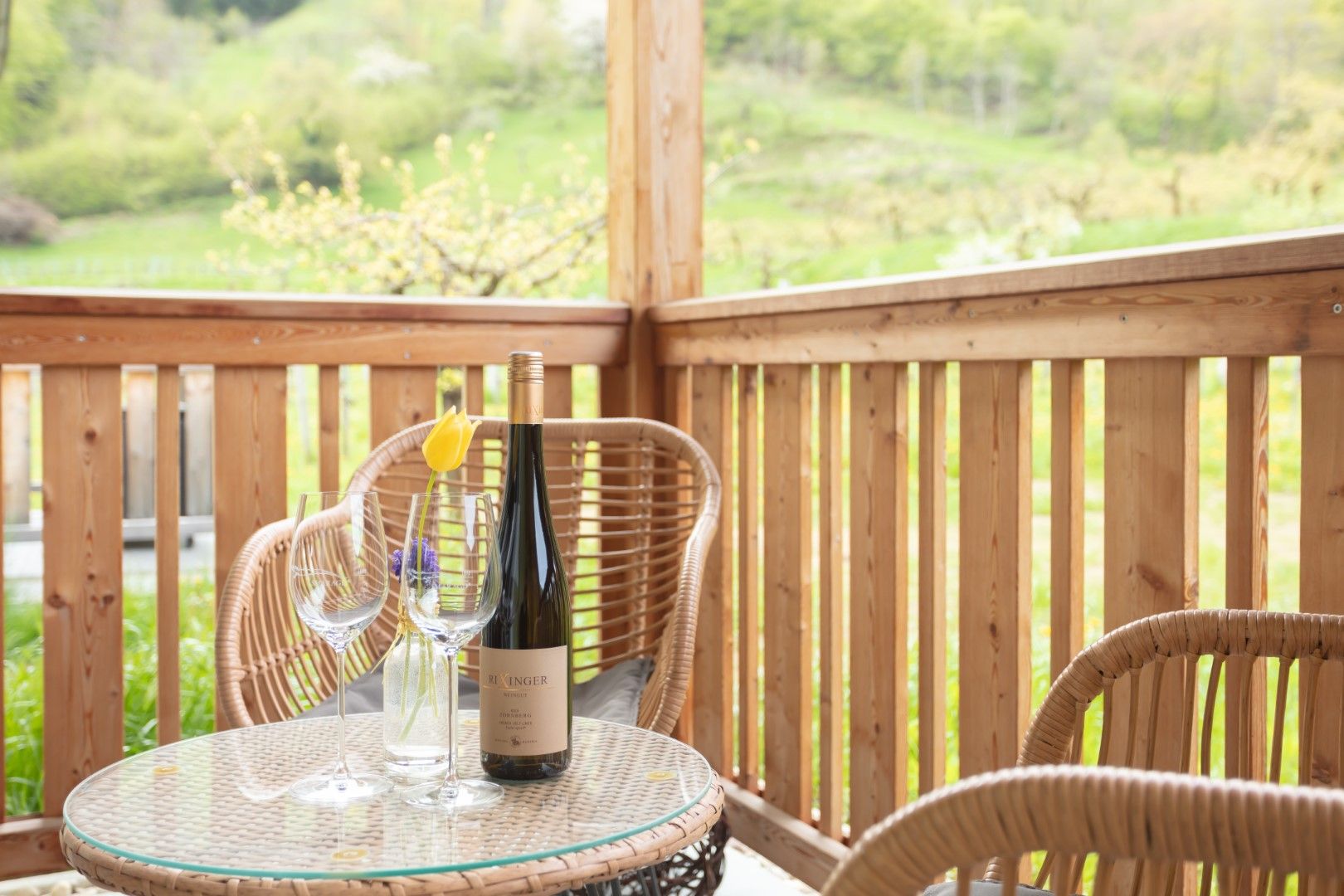 Ein Balkon mit Holzmöbeln, einer Weinflasche und Gläsern auf einem Tisch. Im Hintergrund grüne Landschaft.