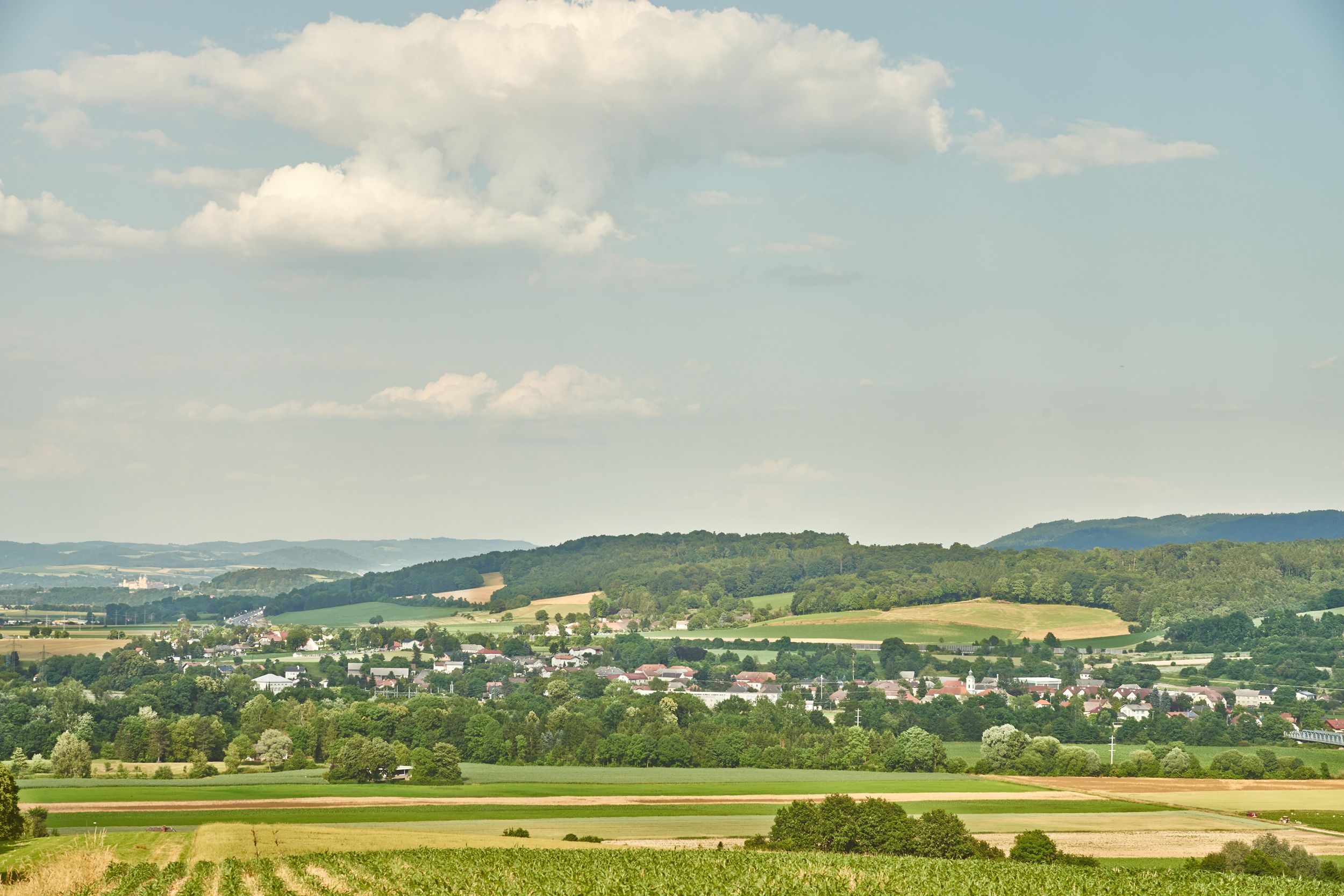 Landschaft mit Feldern, Hügeln und einem Dorf im Hintergrund unter einem bewölkten Himmel.