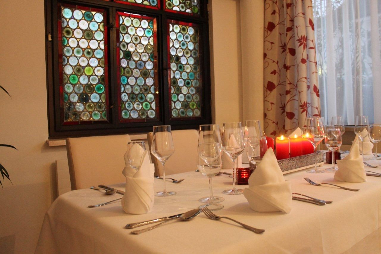 Eleganter Tisch mit weißen Tischdecken, Weingläsern und roten Kerzen vor einem bunten Fenster.