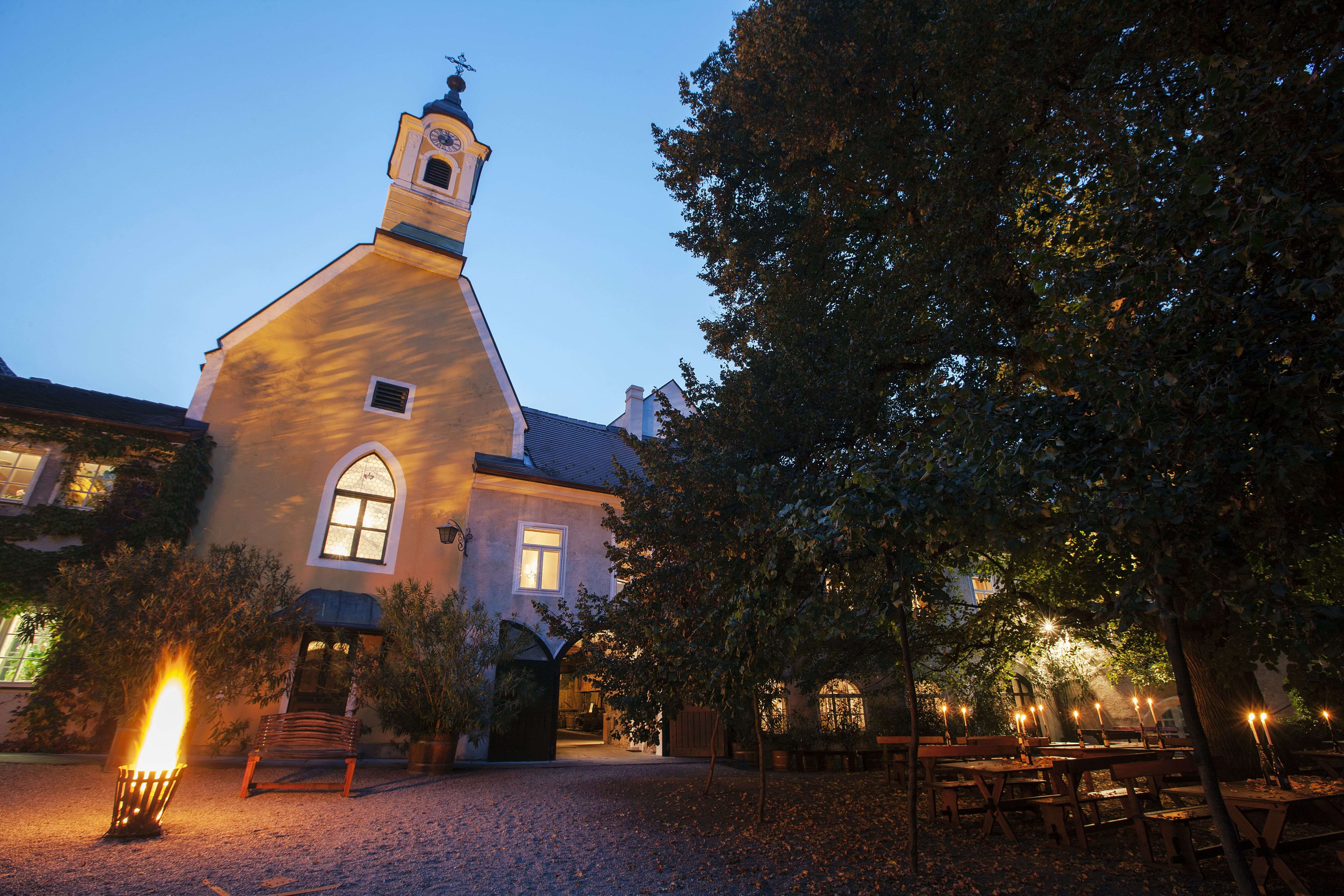 Ein beleuchteter Hof mit einer Kapelle im Abendlicht, umgeben von Bäumen und Tischen mit Kerzen.