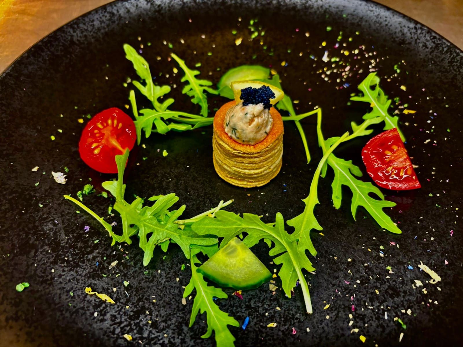 An artfully arranged plate with a small pile of blinis, garnished with caviar, surrounded by rocket, tomatoes and cucumber on a black plate.