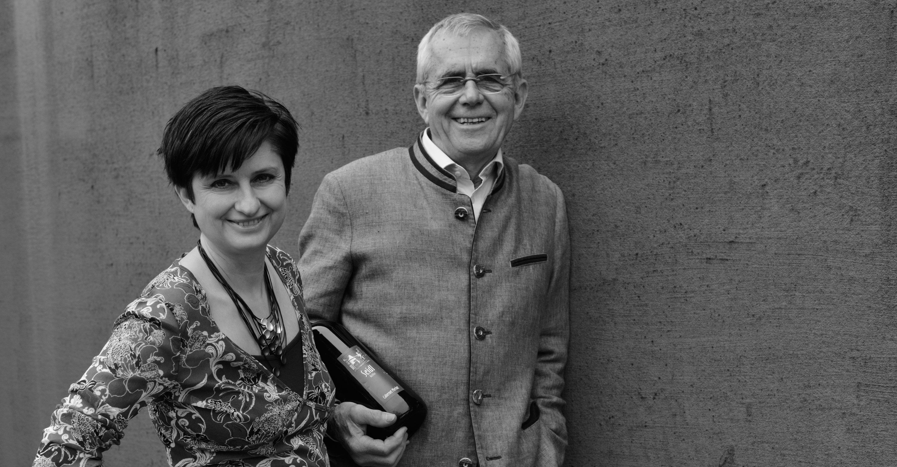 Ein Mann und eine Frau stehen lächelnd vor einer Wand. Der Mann hält eine Weinflasche.