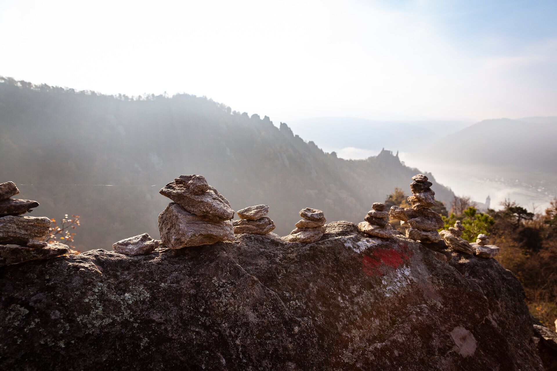 Die sanften Hügel der Wachau erstrahlen im warmen Licht des Herbstes, während die Nebel über dem Tal sanft verwehen. Kleine Steinmännchen säumen den Weg und laden Wanderer ein, die atemberaubende Aussicht auf die umliegende Landschaft zu genießen.