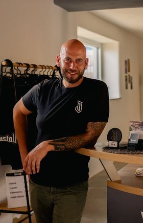 Mann hinter der Theke im Braustall, umgeben von Produkten, Gebäck und Merchandise.