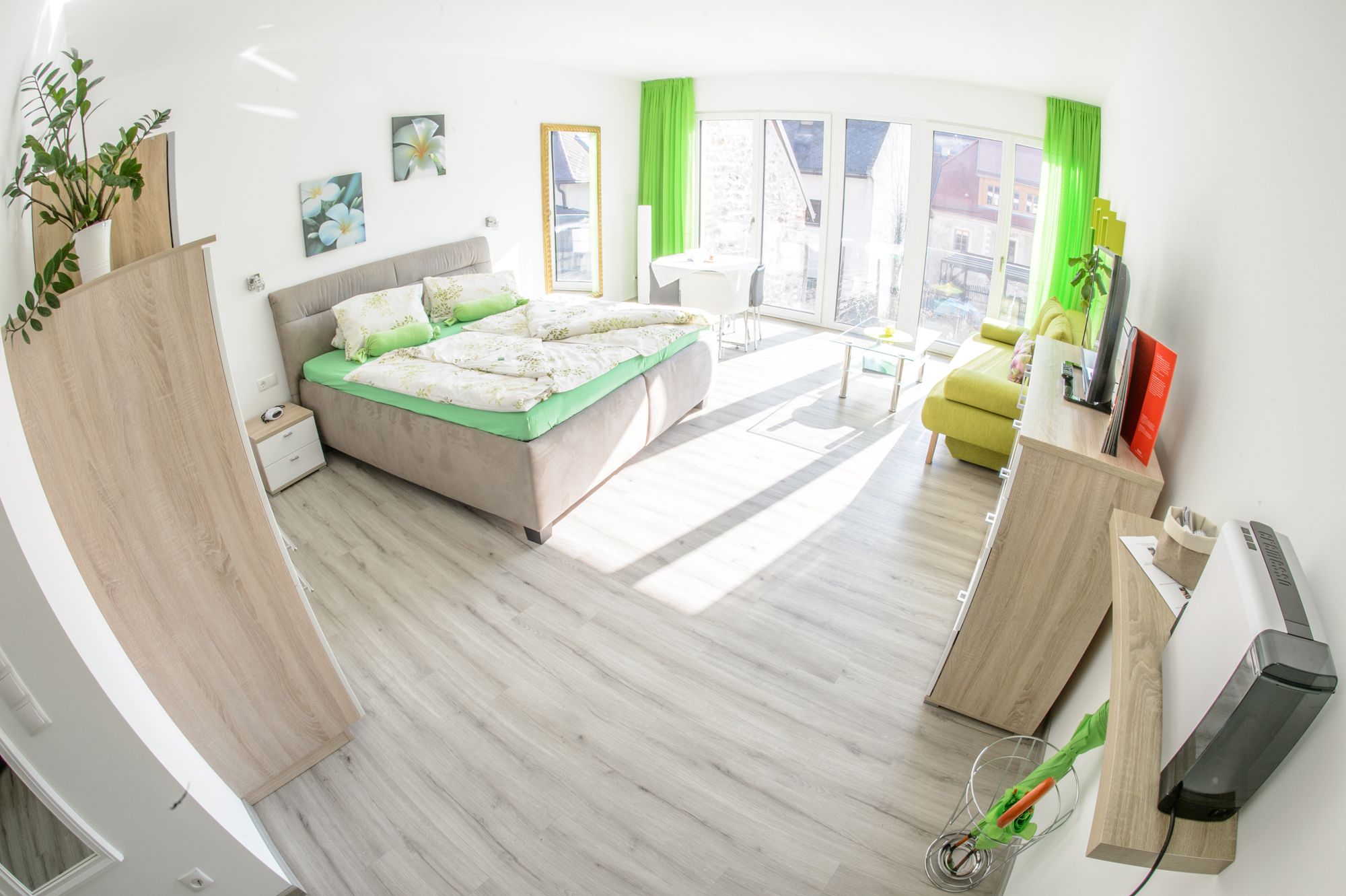 Helles Gästezimmer mit Doppelbett, Sofa, Fernseher und großen Fenstern.