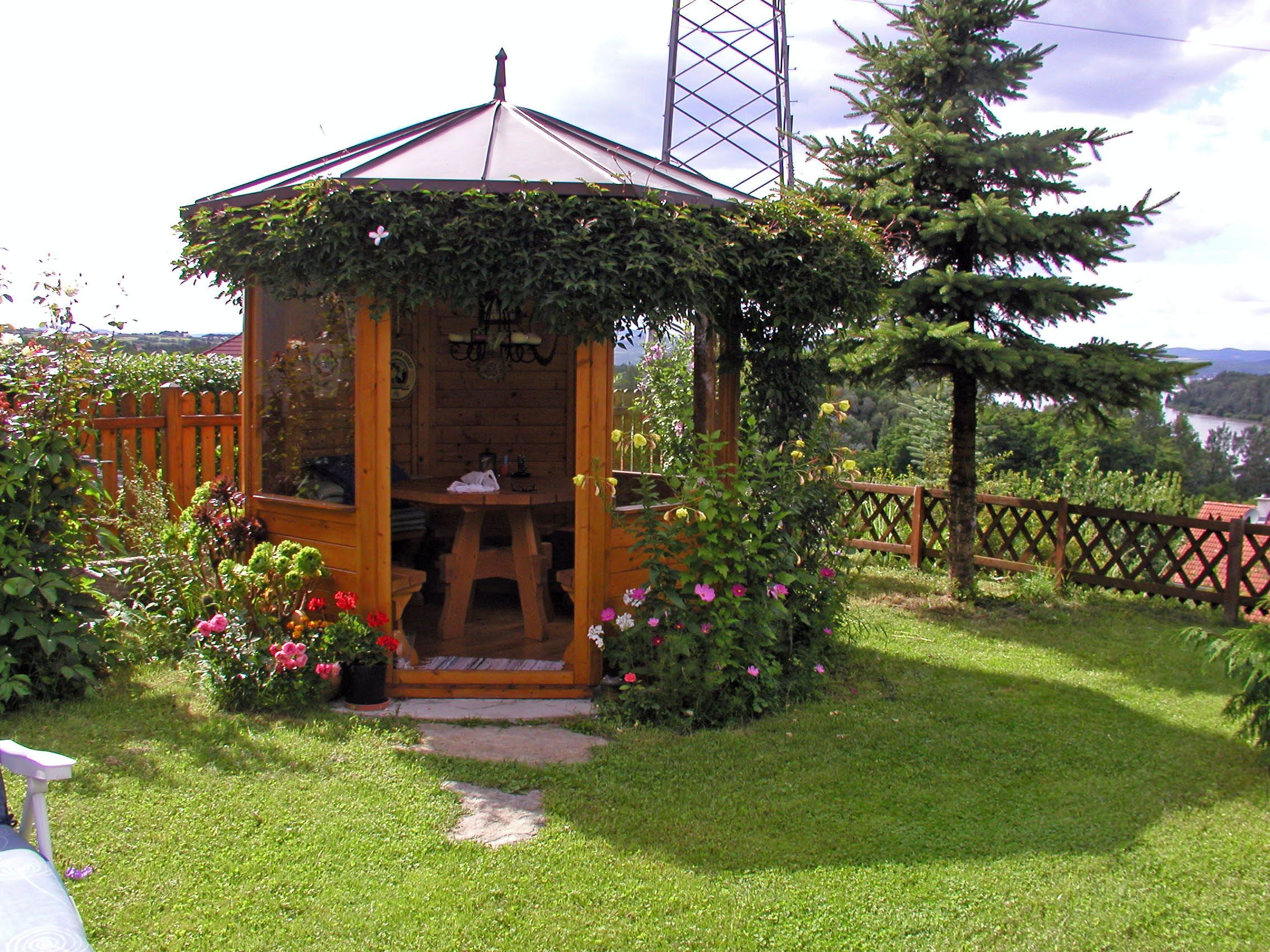 Drevený pavilón v záhrade, obklopený kvetmi a stromami, s výhľadom na jazero v pozadí.